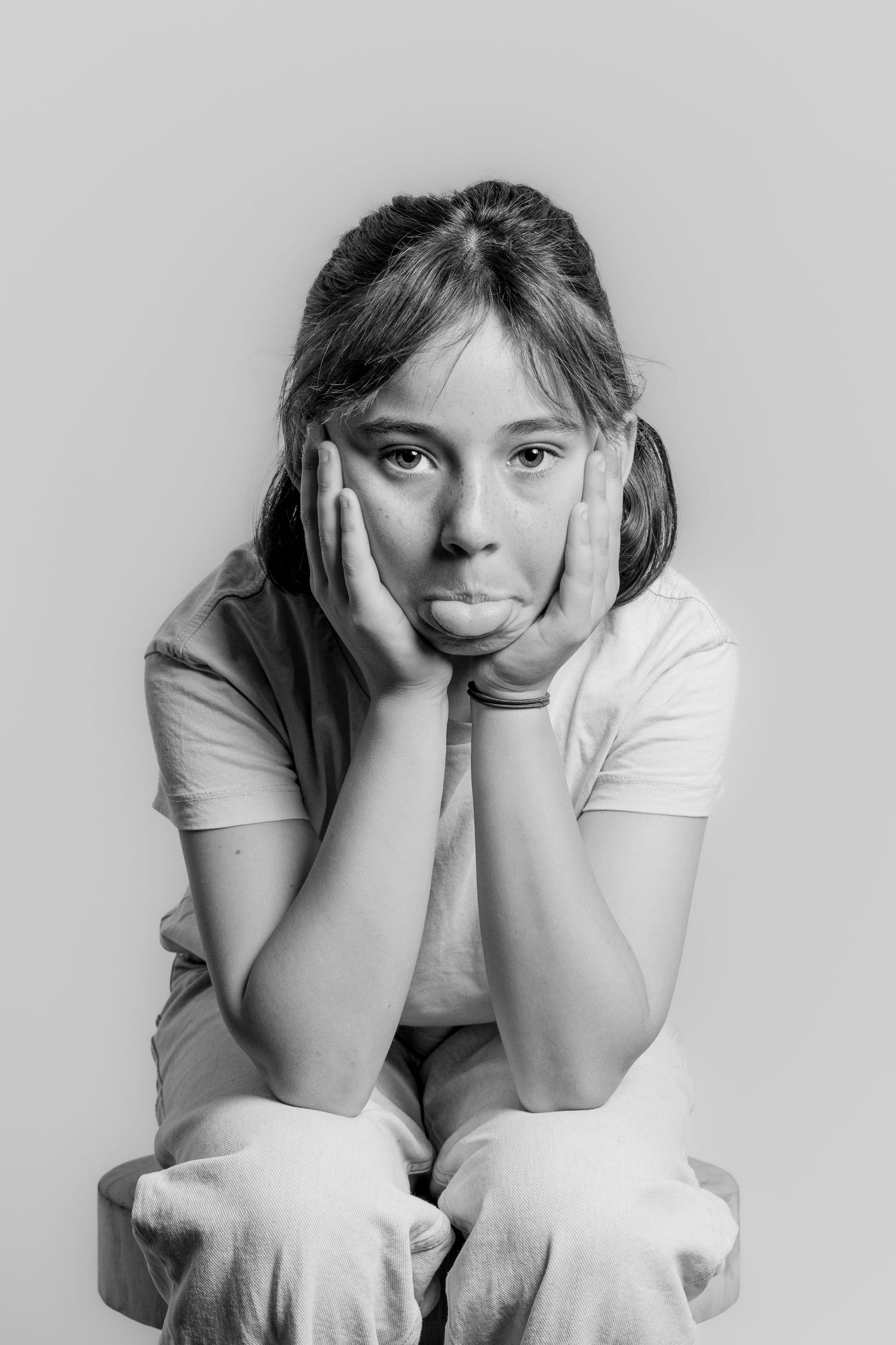A young girl makes a pouty face with hands on cheeks in a black and white portrait.
