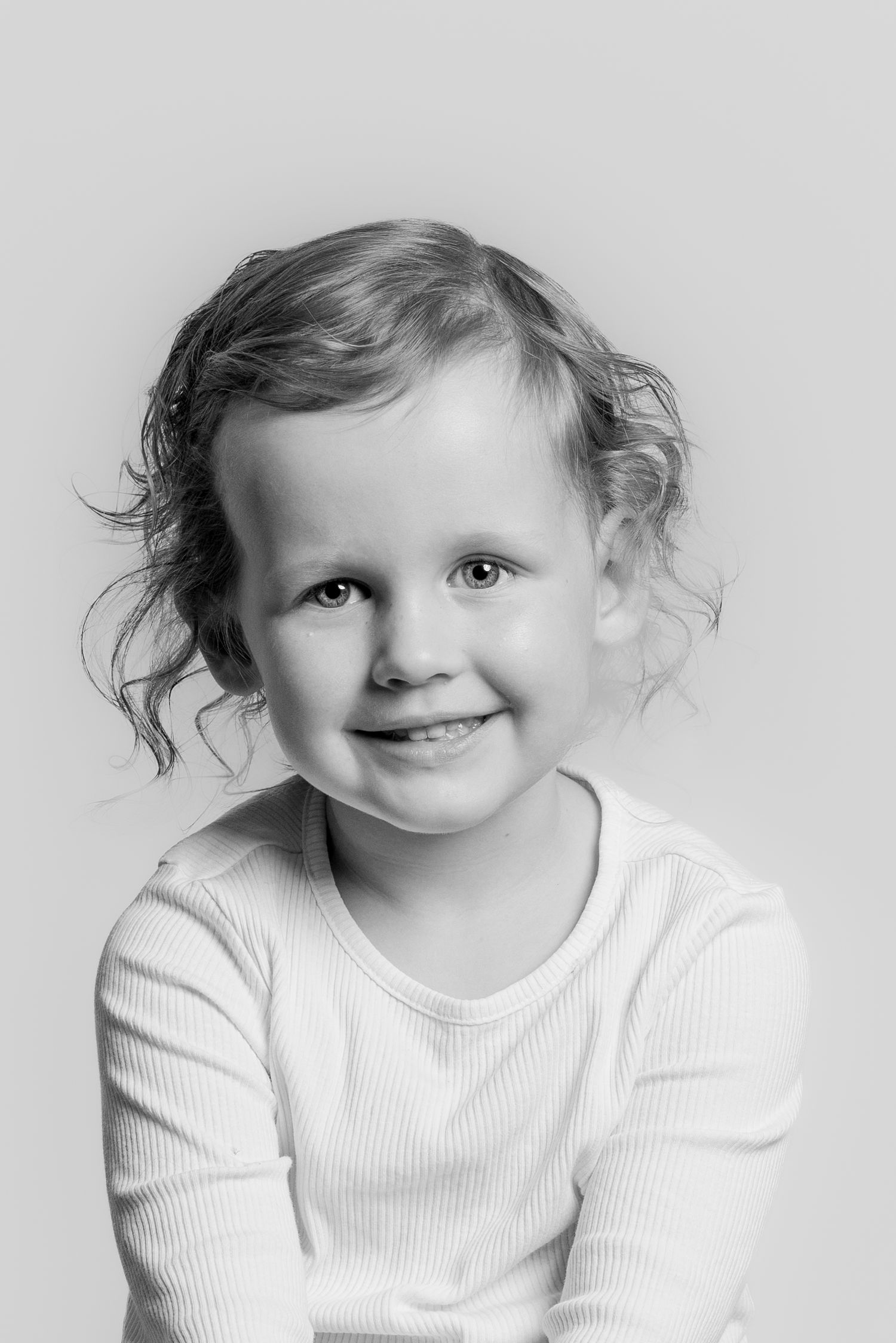 Black and white studio portrait of a smiling young girl with curly hair in a white ribbed top.