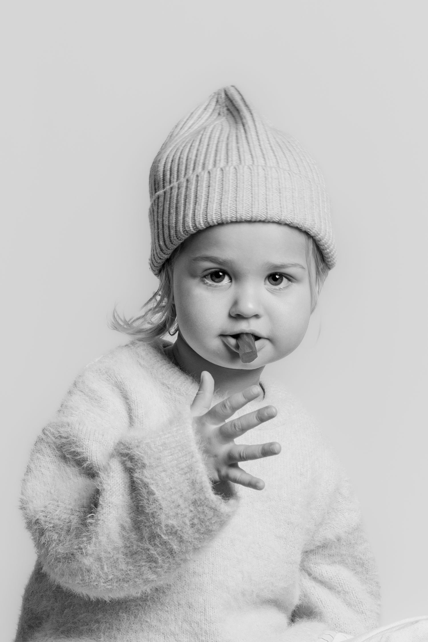 Black and white portrait of a toddler in a knit hat and fuzzy sweater, sticking out tongue.