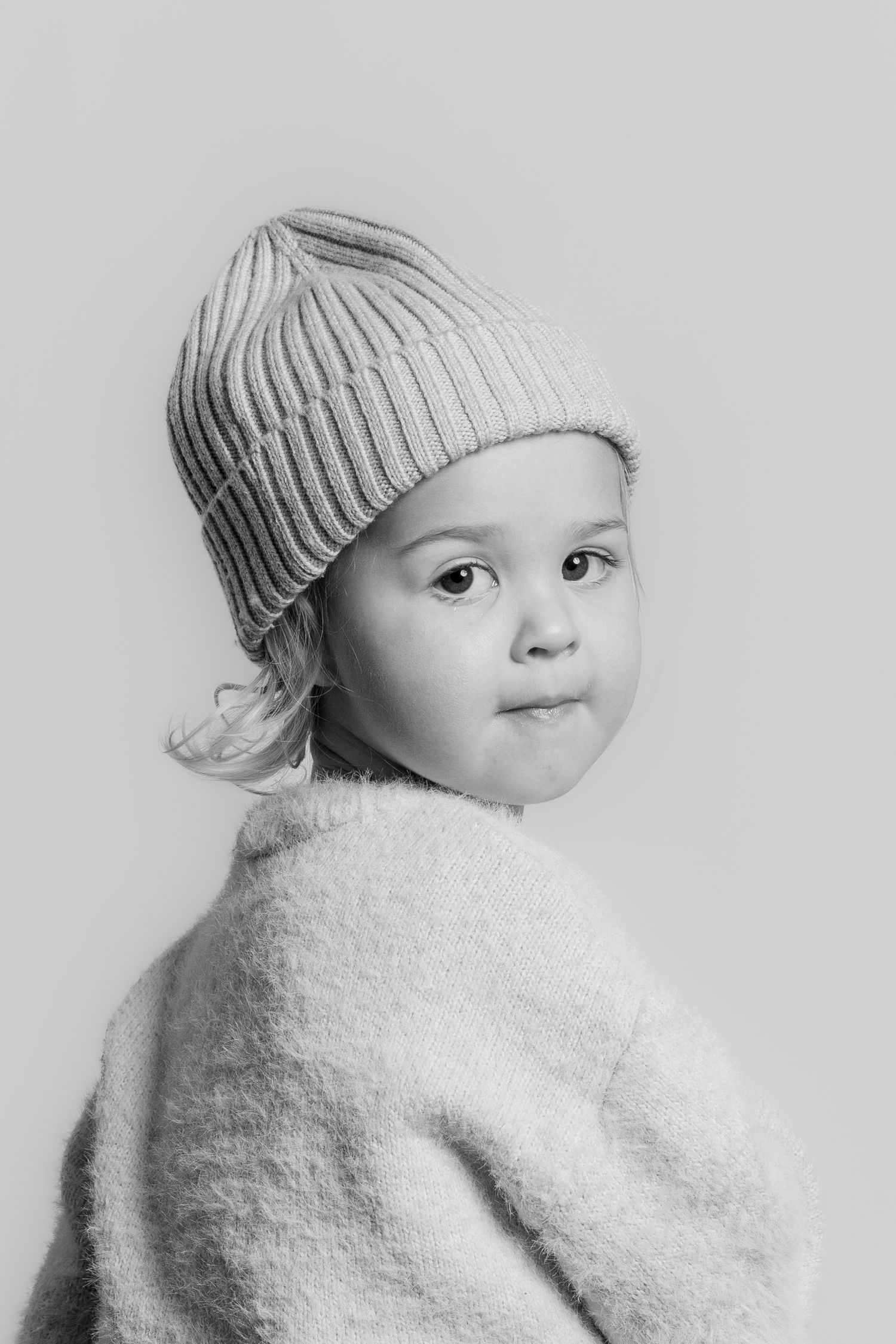 Black and white studio portrait of a young child in a ribbed knit beanie and cozy sweater looking over their shoulder.