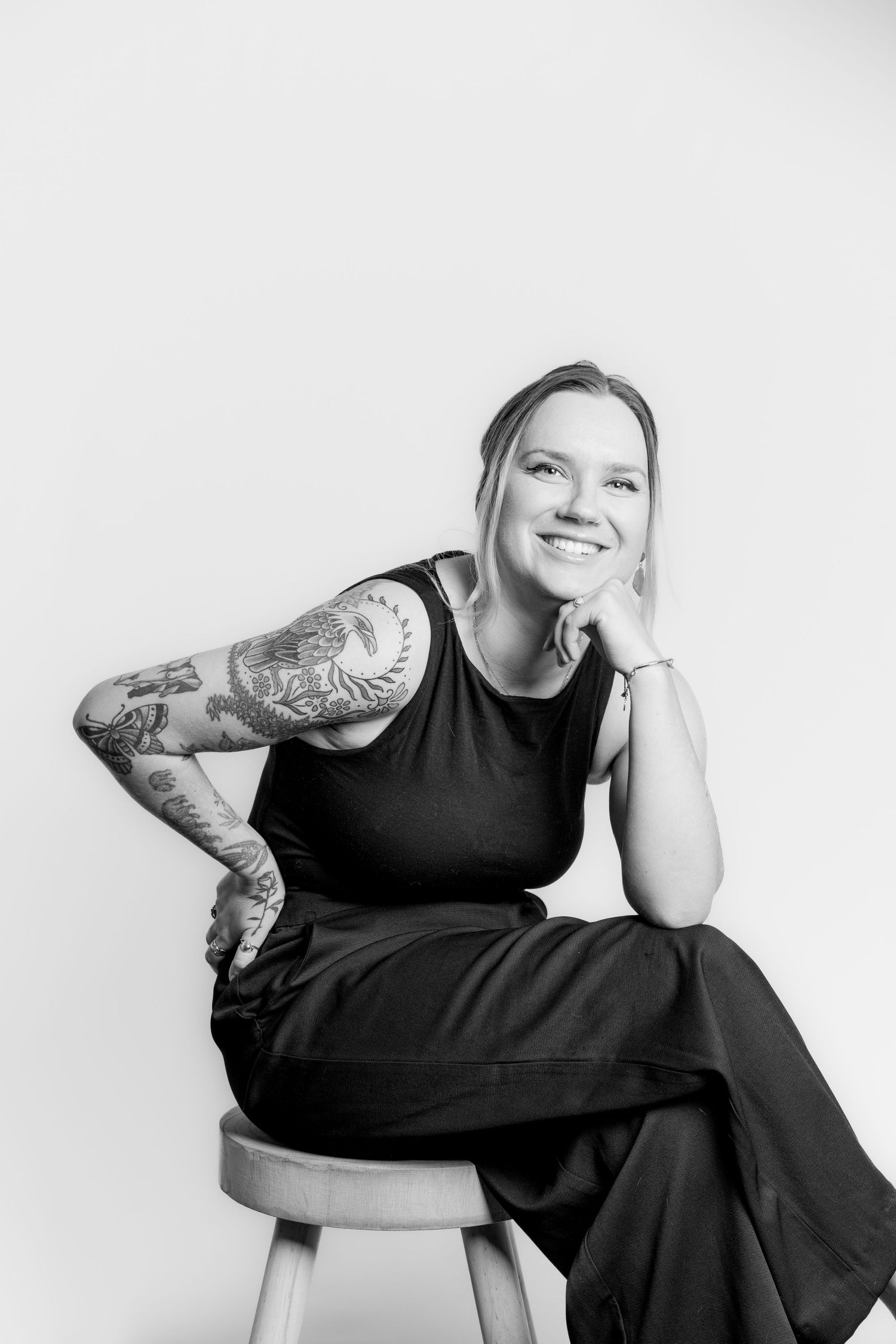 Black and white portrait of laughing tattooed blonde woman seated on stool in studio.