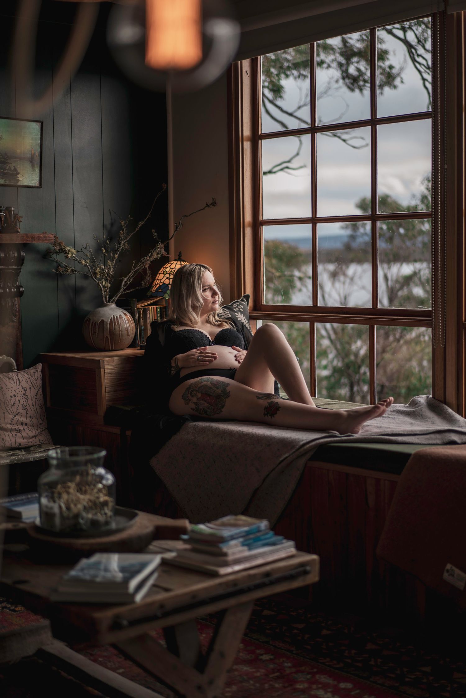Tattooed pregnant woman sitting peacefully by a large window in a warmly lit room.