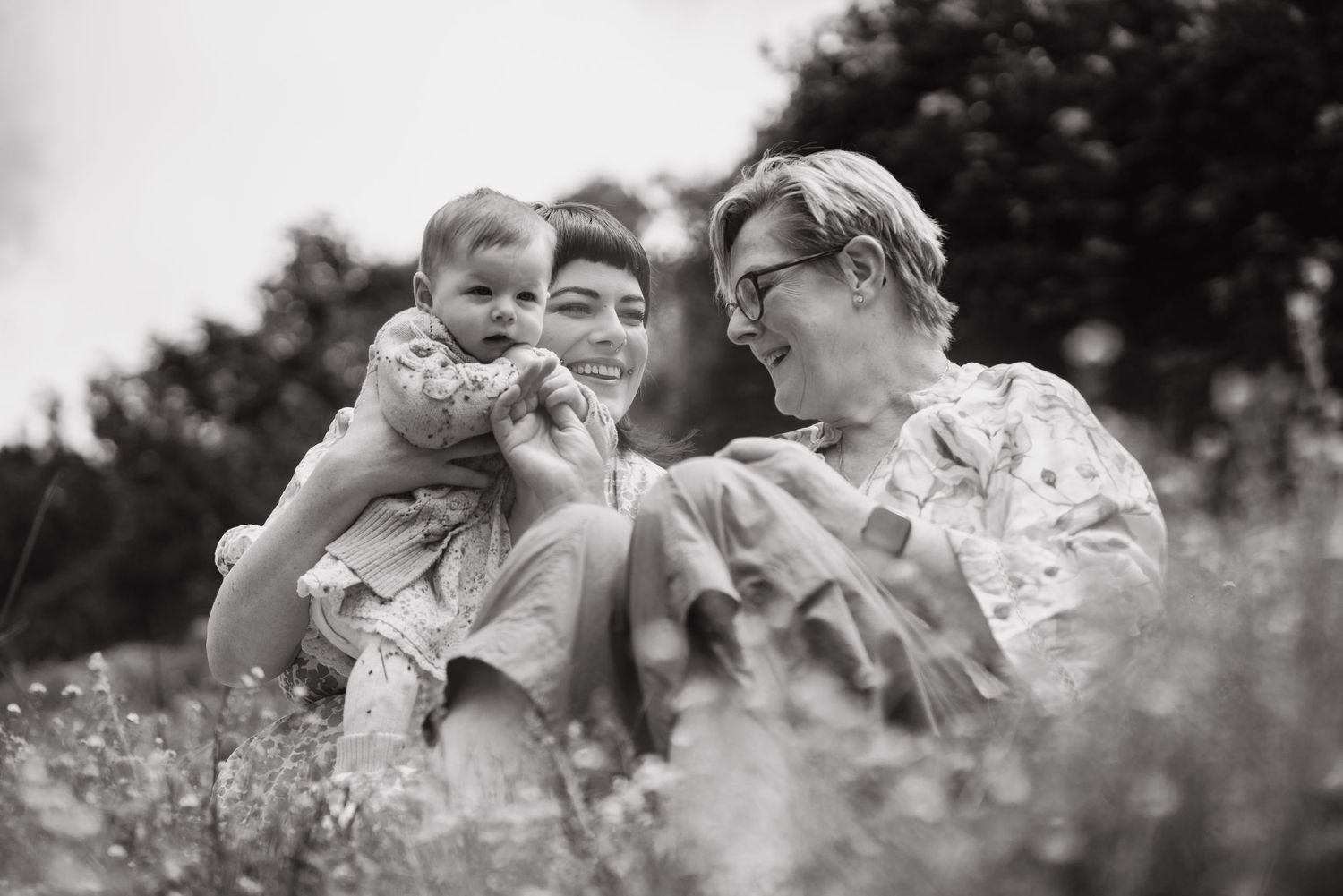 Black and white photo of two mothers laughing with their baby in a meadow.