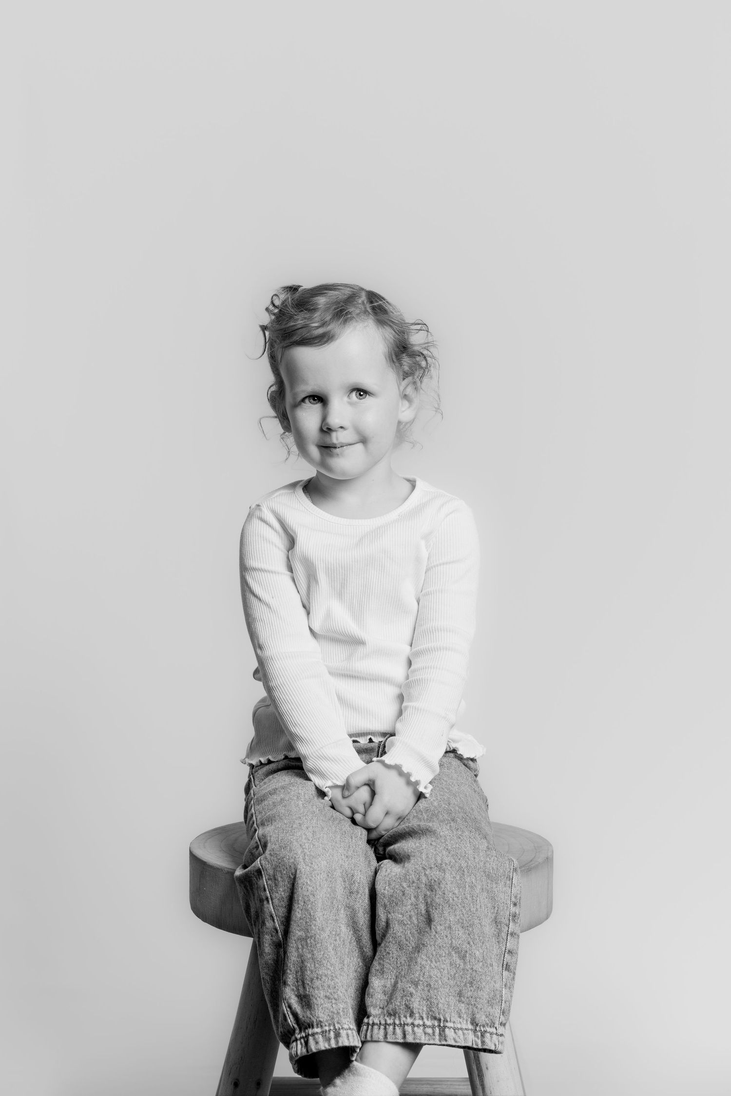 Black and white portrait of a young girl with curly hair sitting on a wooden stool, smiling.