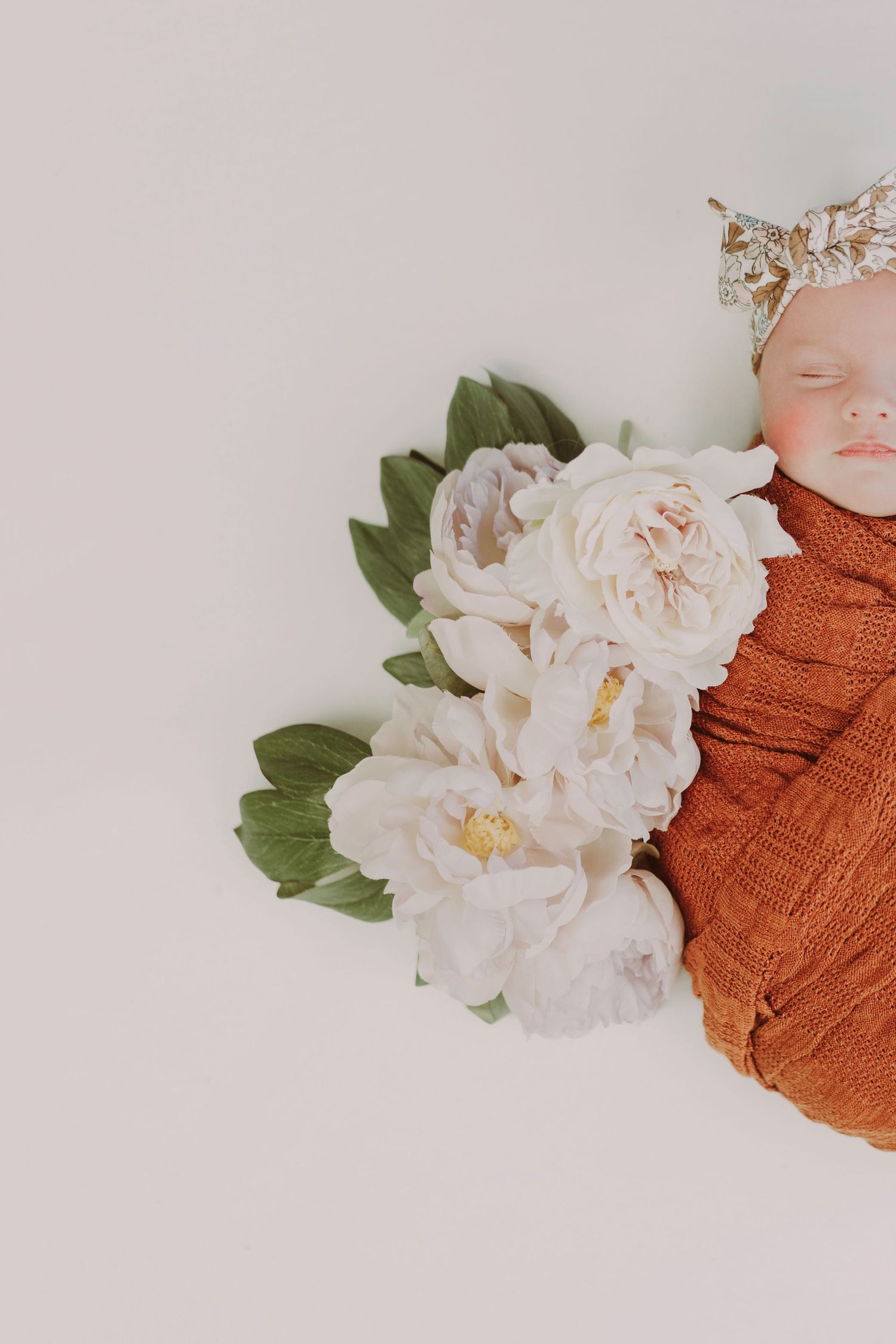 Sleeping newborn baby swaddled in rust orange wrap with white peonies and a floral crown on white backdrop.