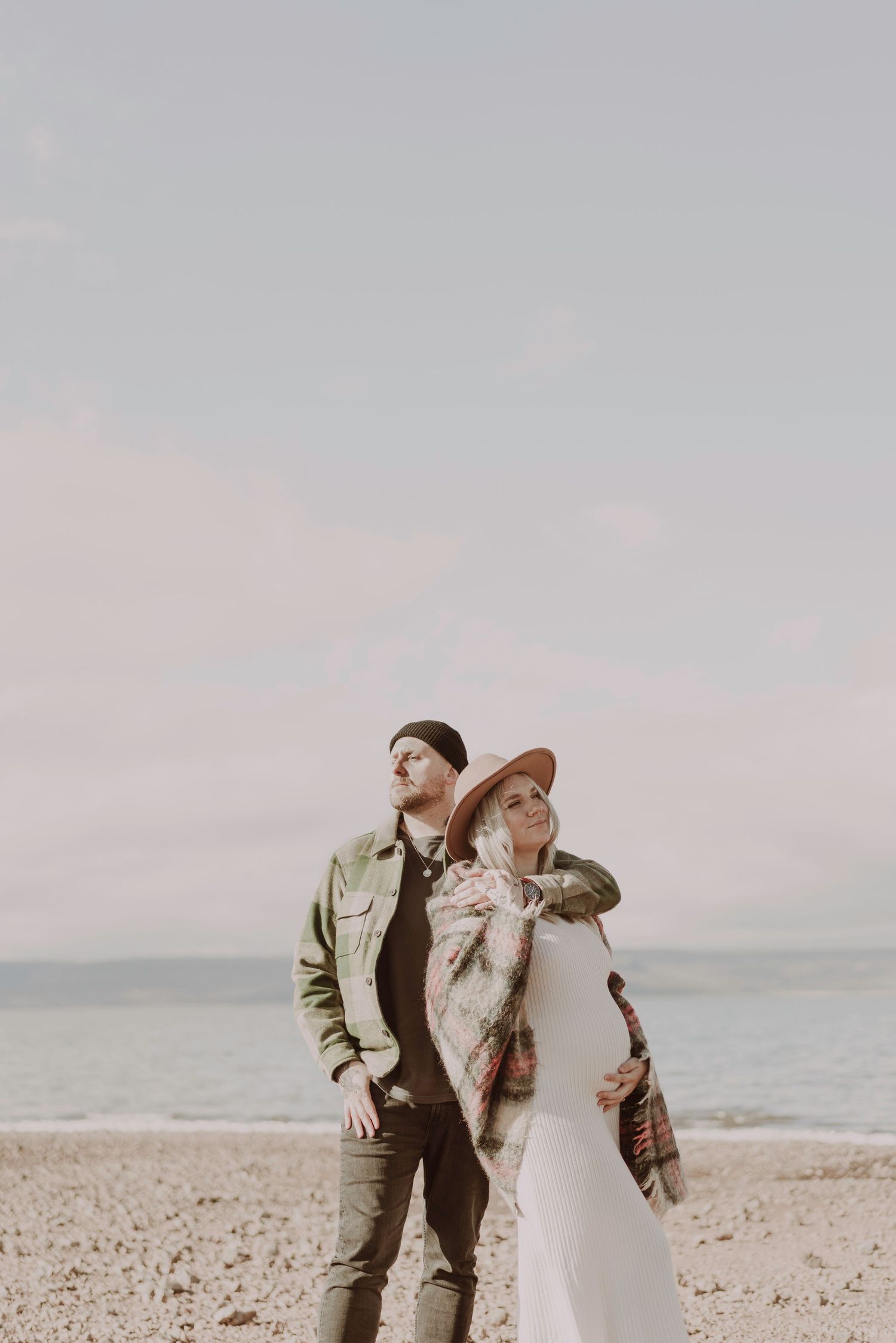 Pregnant couple embracing on a pebble beach under a soft cloudy sky.