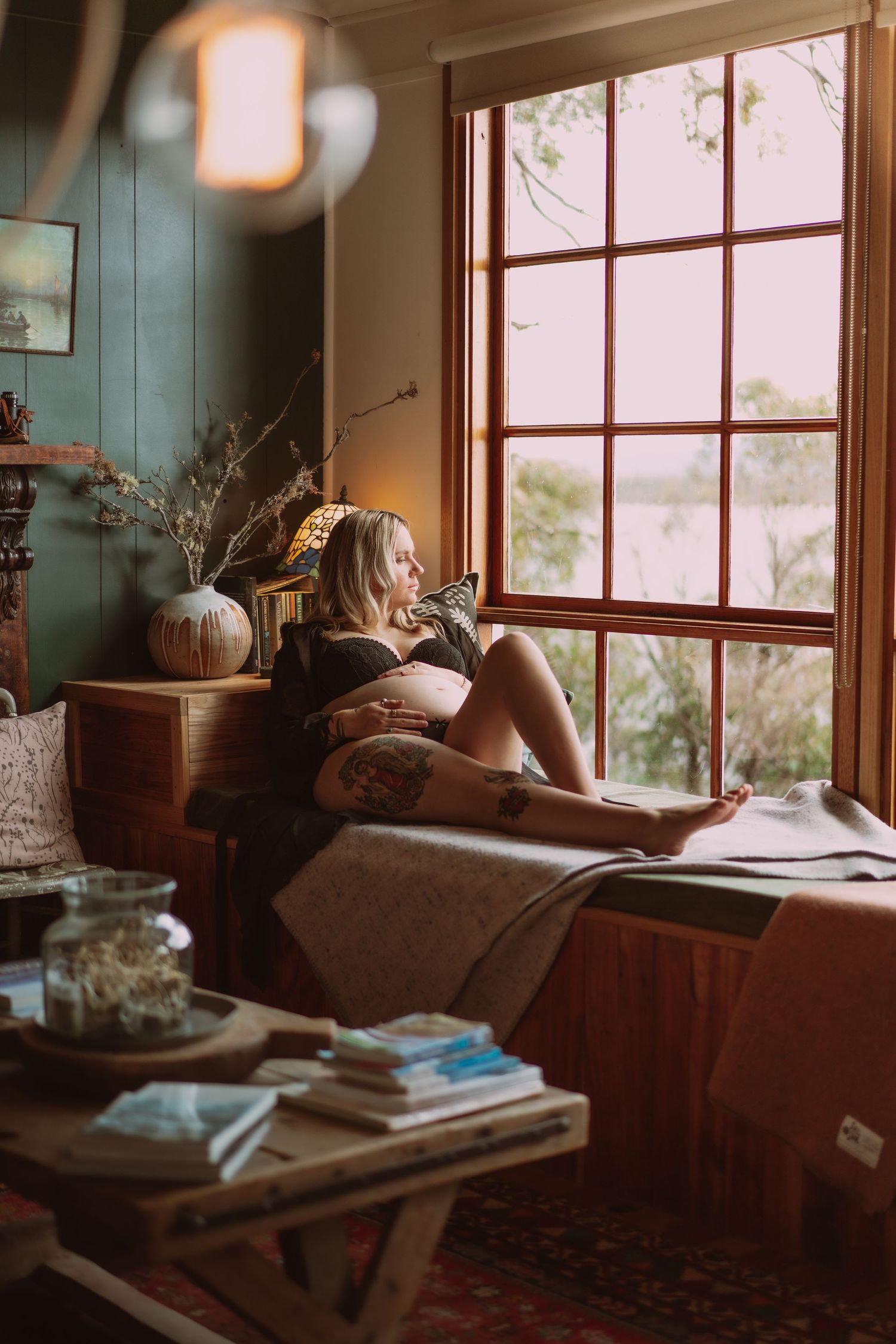 Pregnant woman in lingerie relaxing by a large wooden window in a cozy, warmly lit rustic room. Tasmania Coldwater Cabin Air bnb