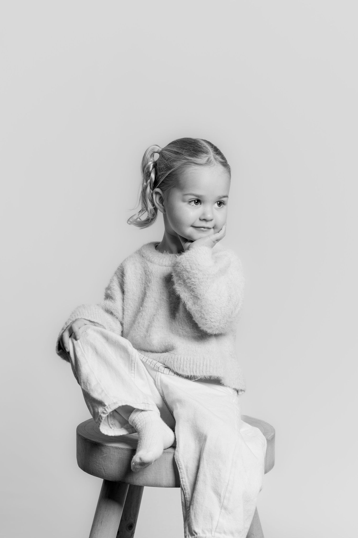 Black and white studio portrait of a young girl with pigtails sitting on a wooden stool in a fluffy knit sweater.