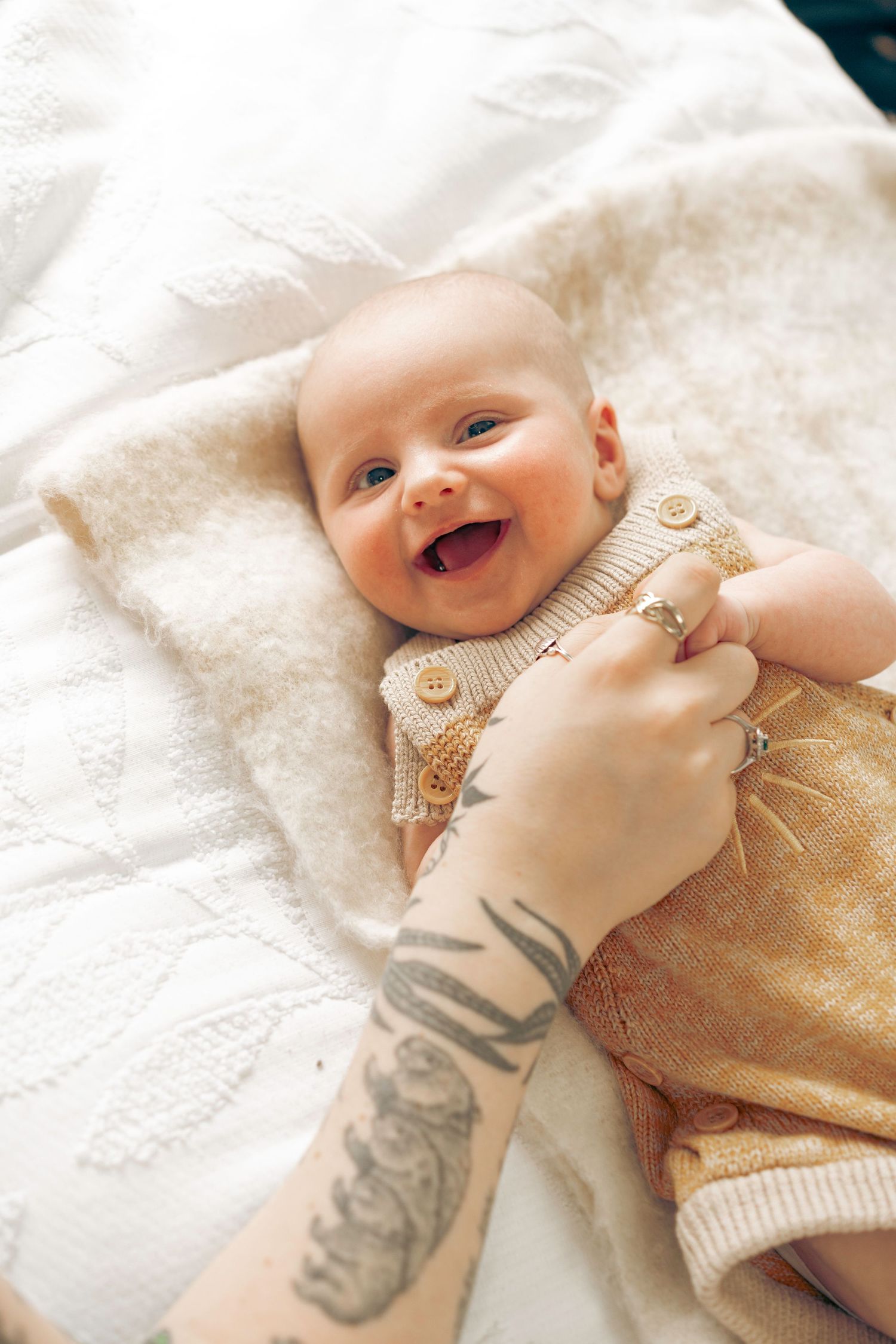 Smiling newborn baby in knit outfit held by tattooed parent on white blanket.