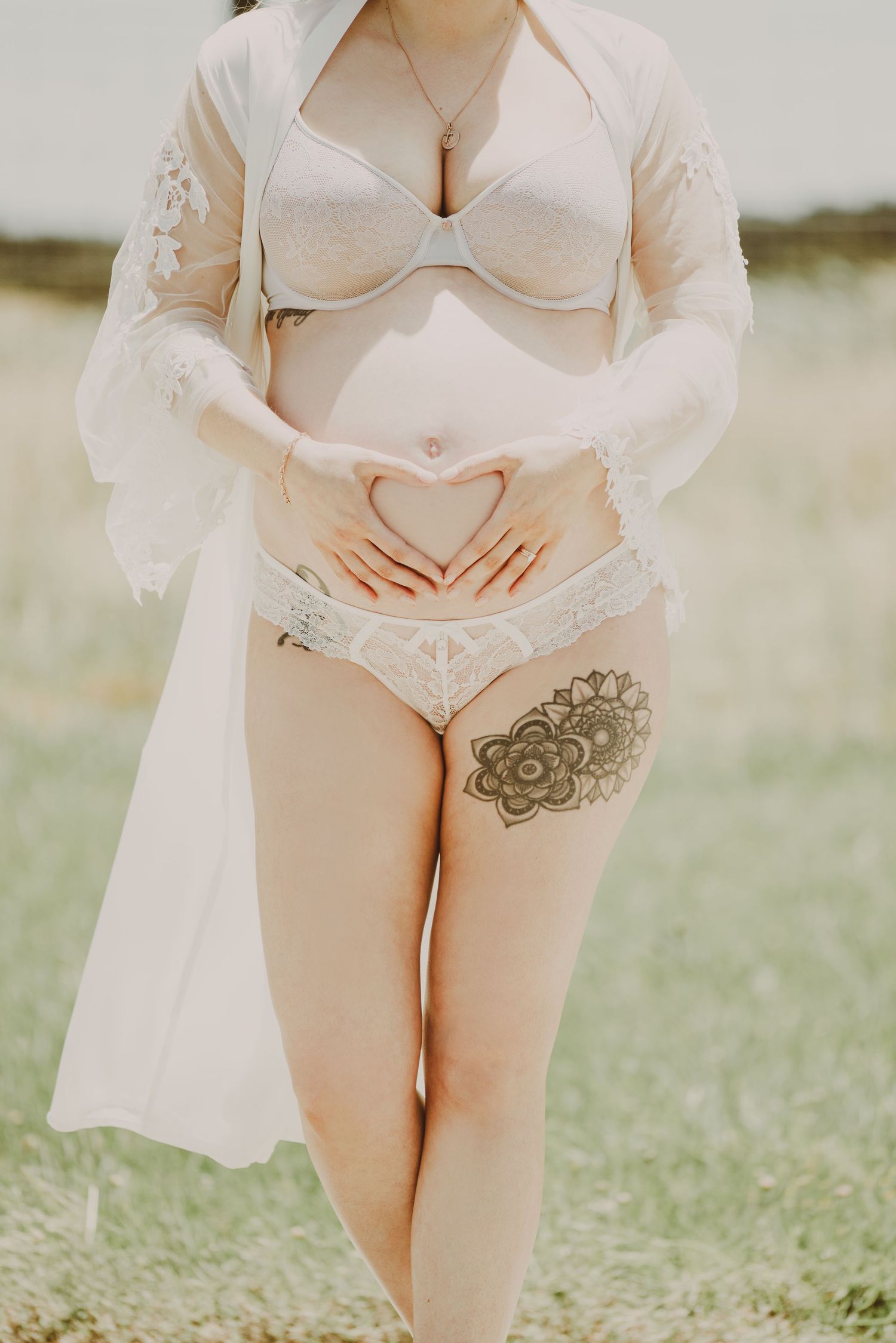 Pregnant woman in white lace lingerie making heart shape with hands over baby bump outdoors.