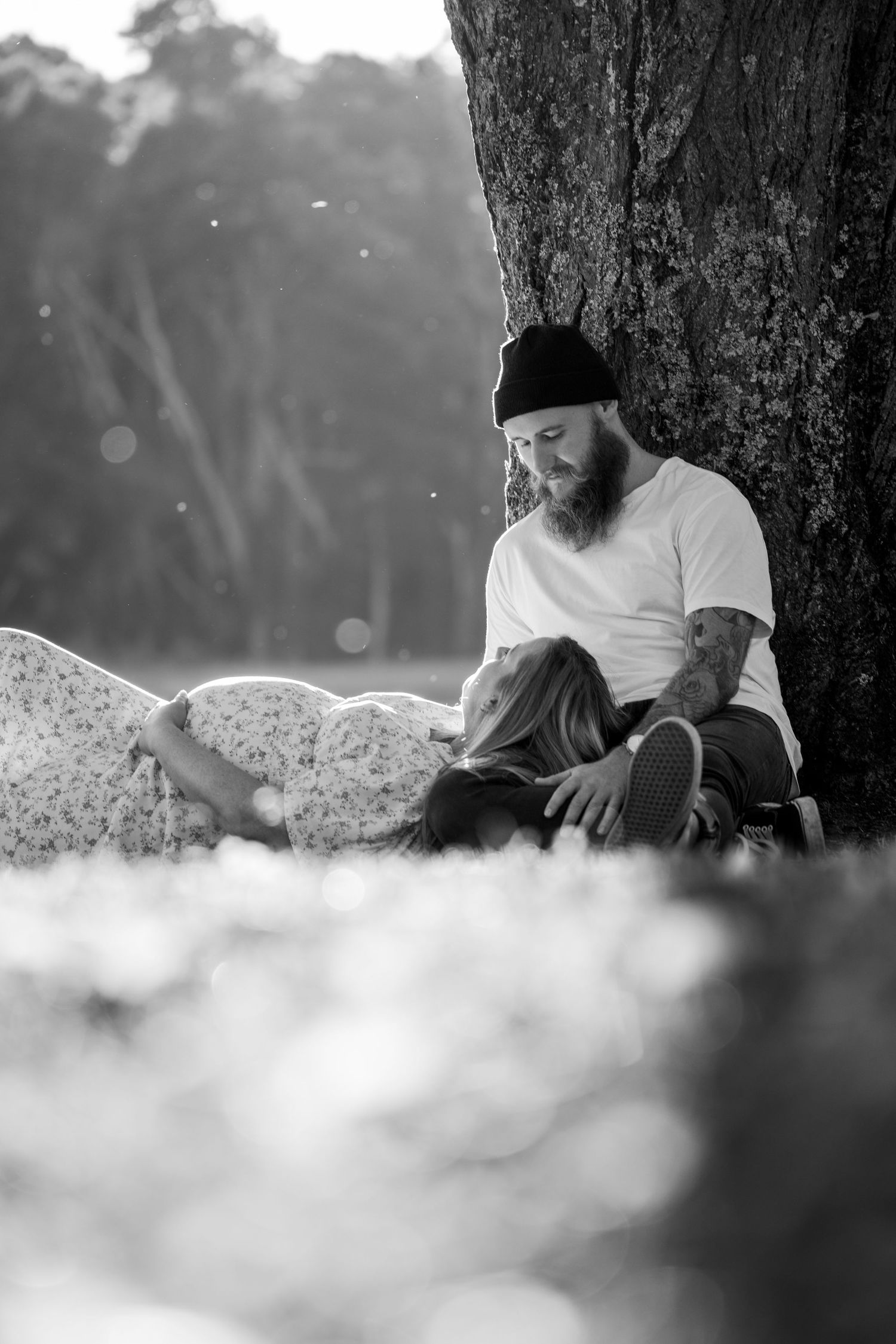 Bearded man in beanie sitting under tree with pregnant partner lying across his lap, black and white.