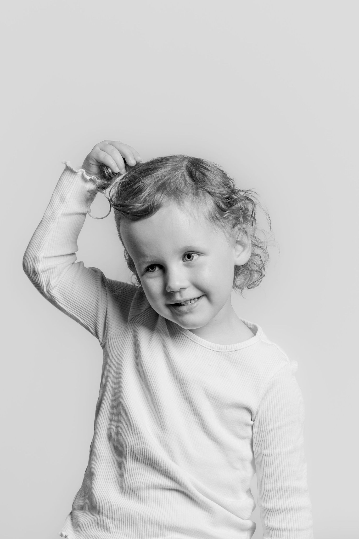Black and white portrait of a playful toddler girl with curly hair, twirling her hair and smiling.