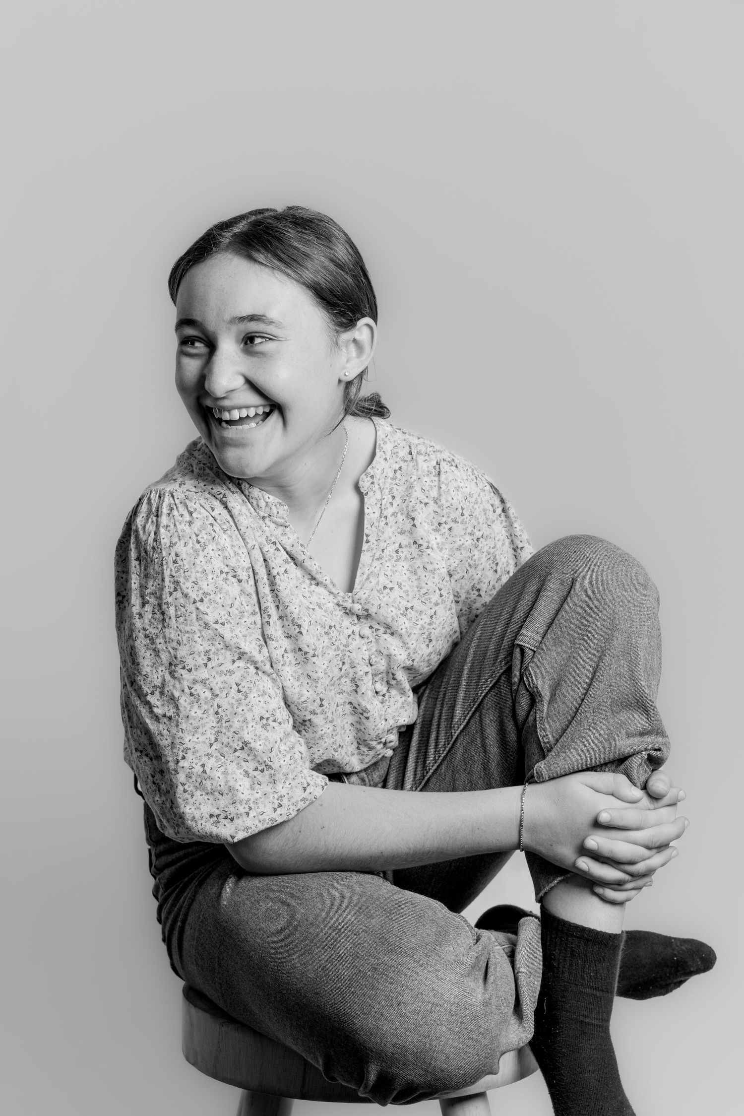 Black and white photo of a laughing teenage girl sitting cross-legged on a stool in a floral blouse.