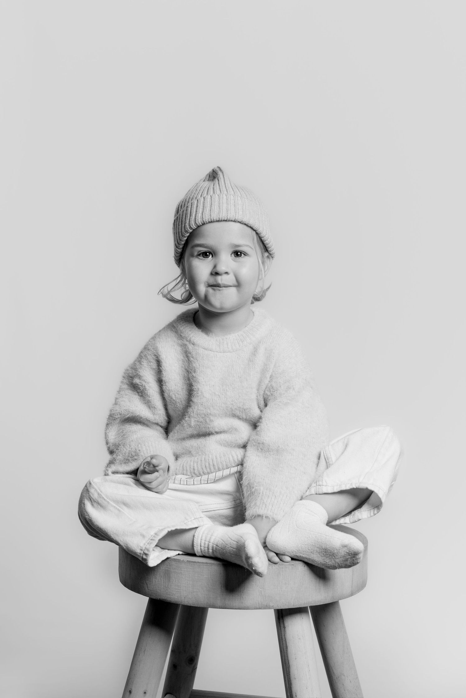 Black and white photo of a smiling toddler wearing a knit hat and sweater sitting cross-legged on a wooden stool.