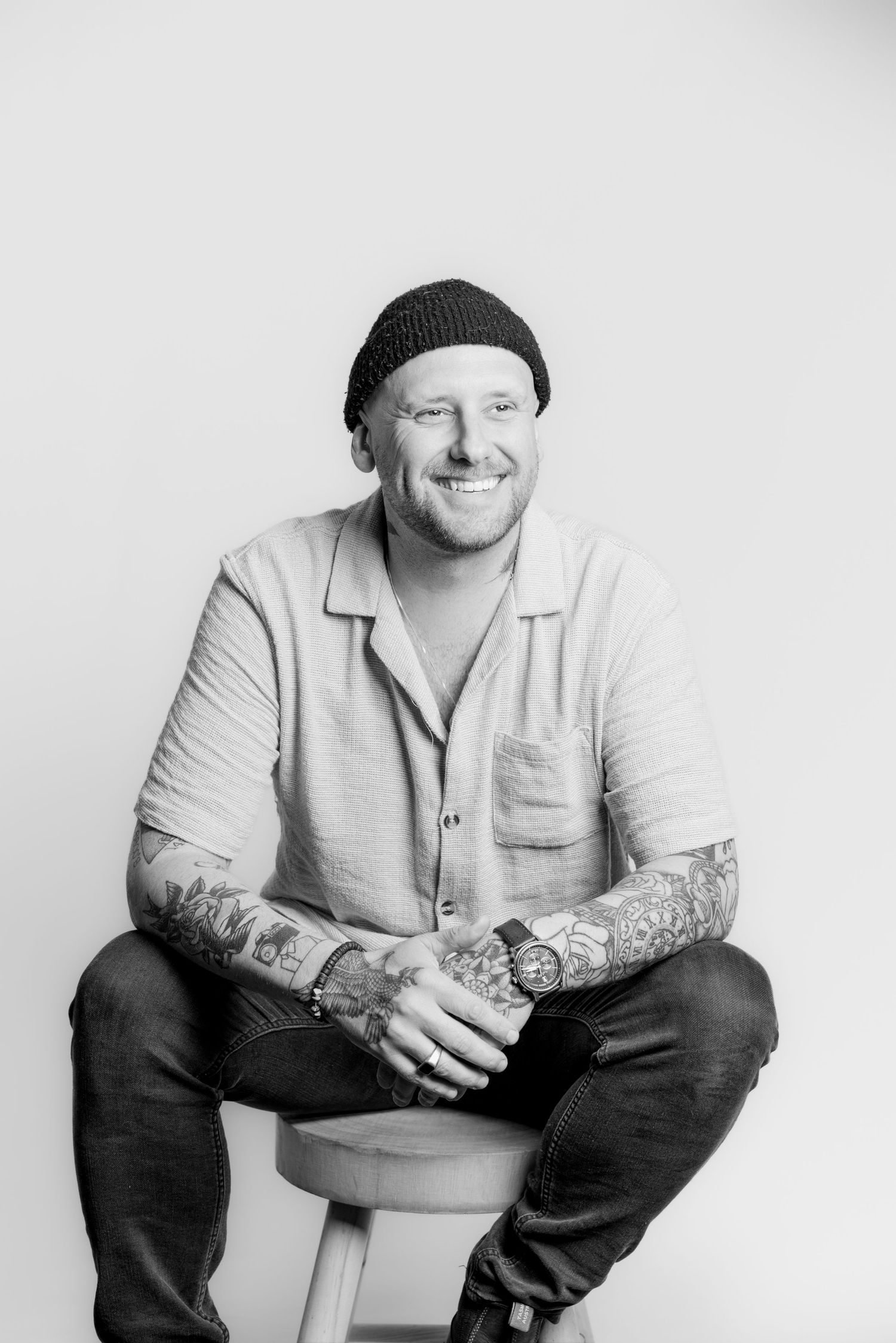 Black and white studio portrait of smiling tattooed man in beanie seated on stool.