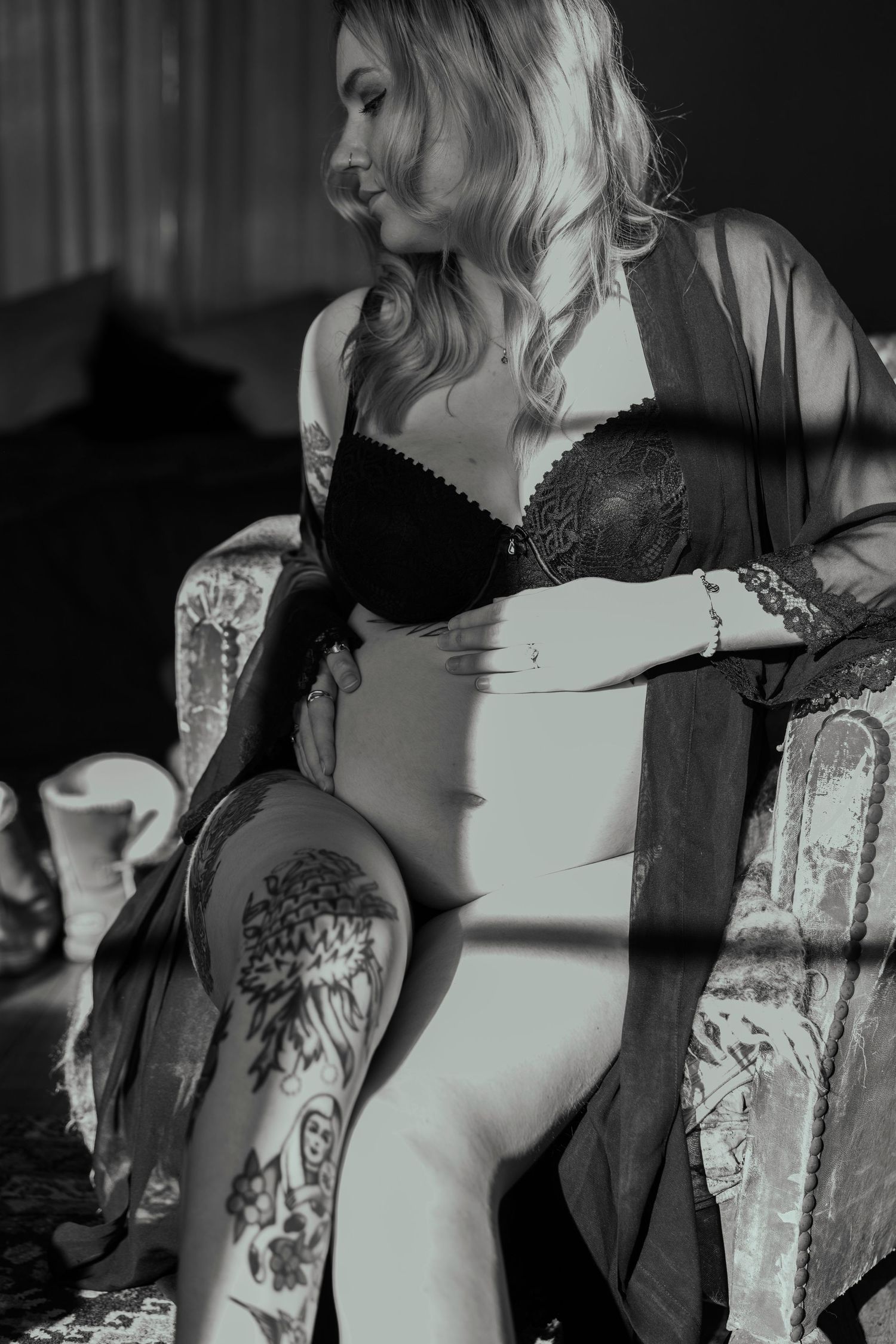 Black and white photo of tattooed pregnant woman in black lingerie sitting in vintage chair.