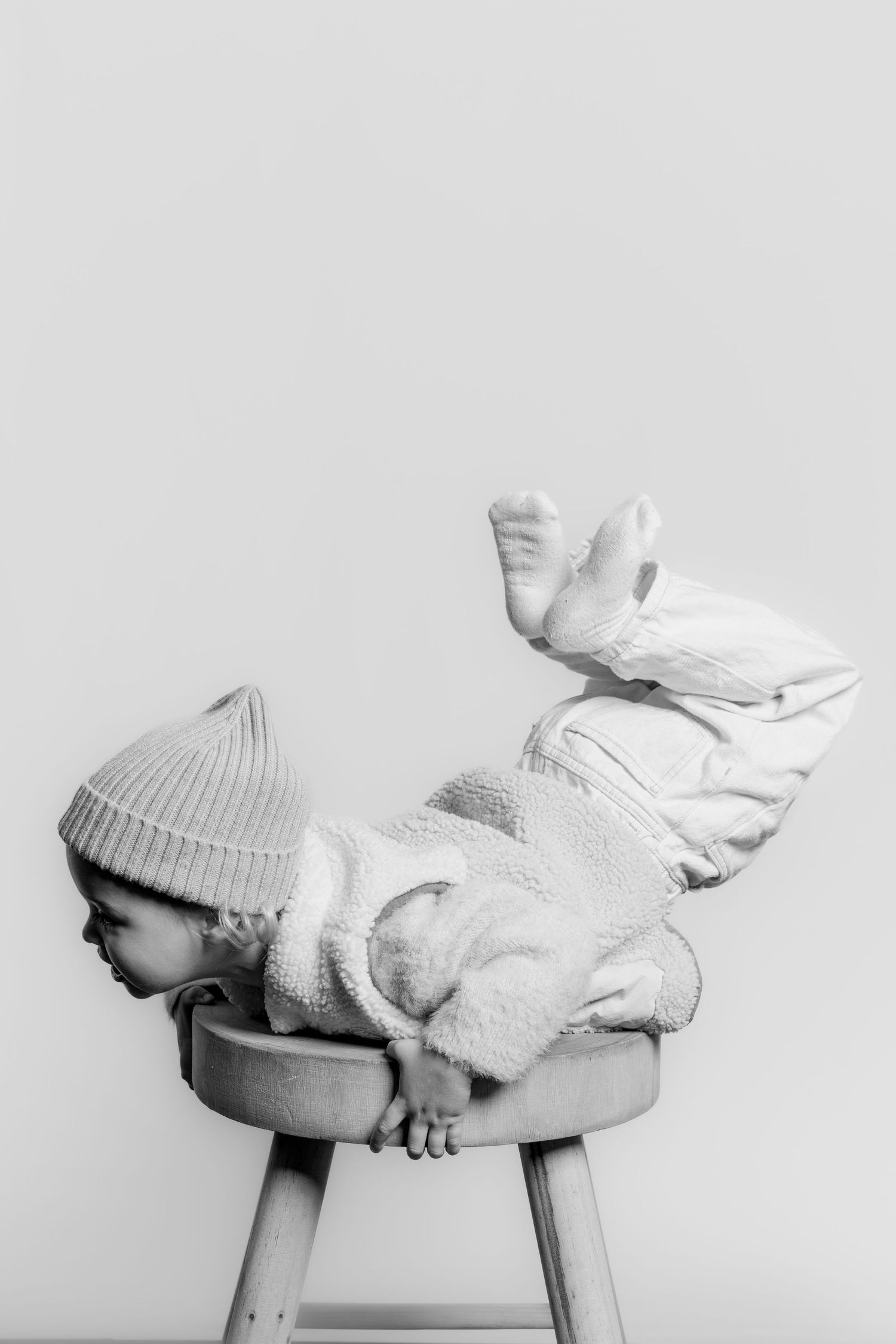 Black and white photo of a playful toddler in a knit beanie dramatically draping themselves over a small wooden stool.