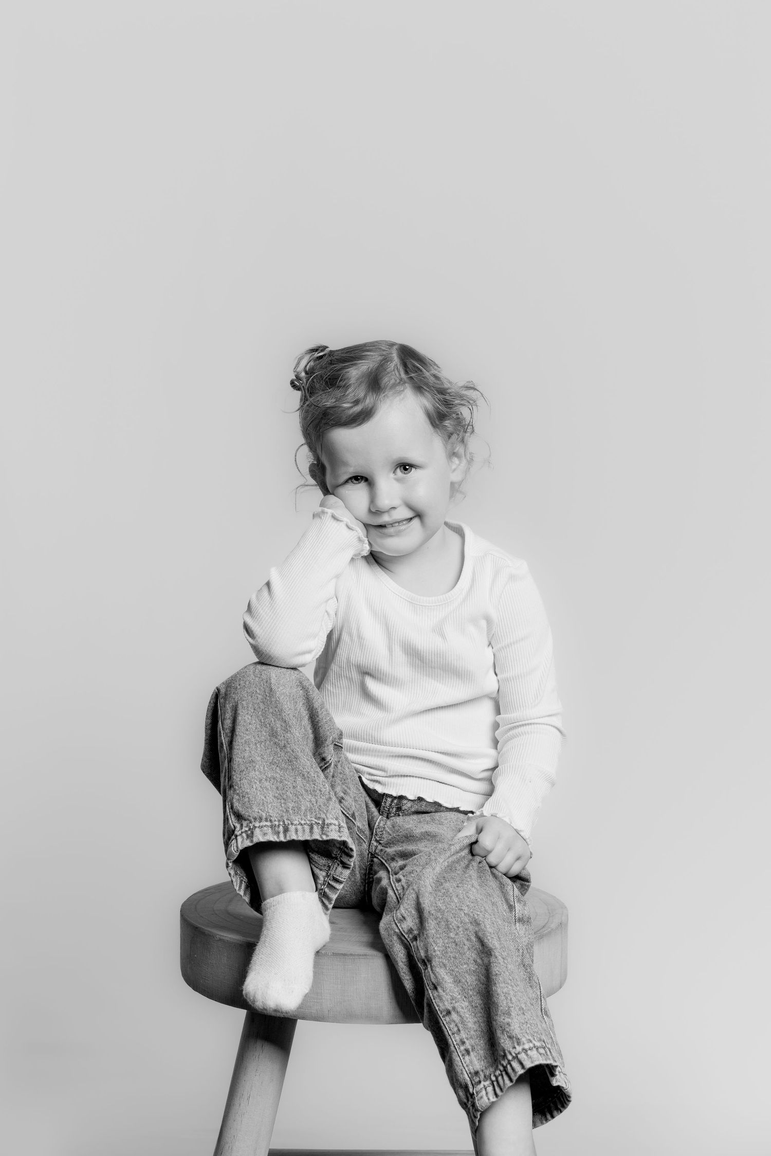 Black and white portrait of a young girl sitting on a stool, smiling with hand on her cheek.