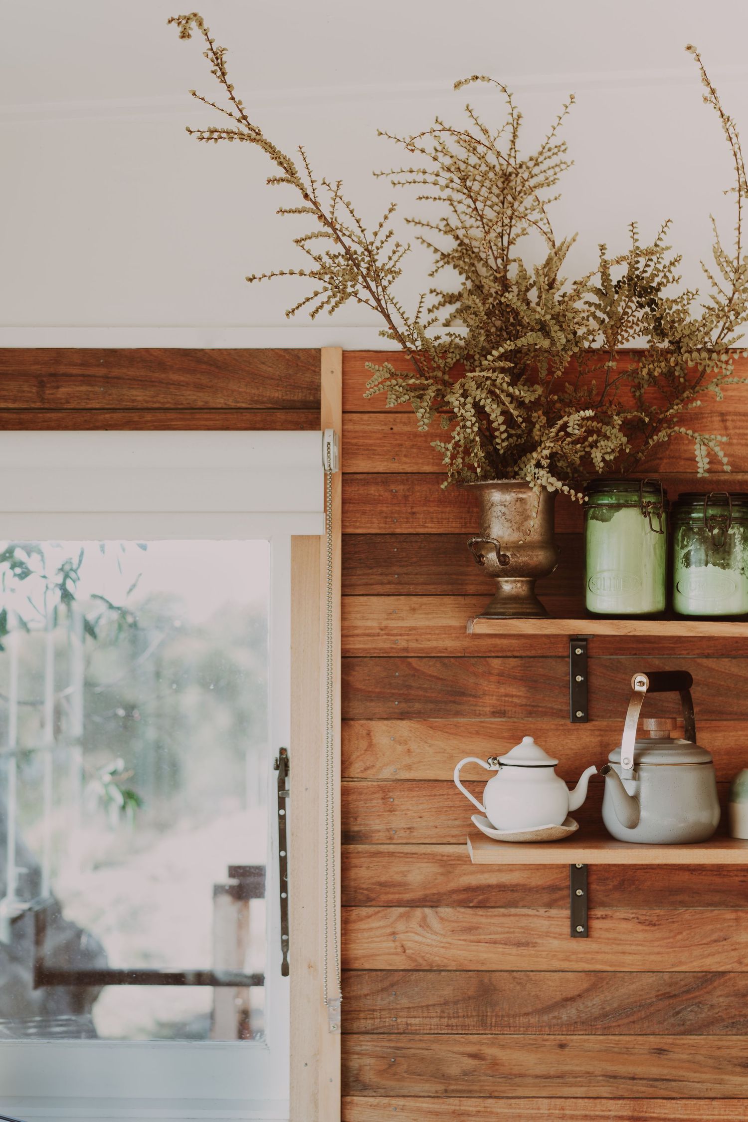 Rustic wooden shelves with white teapot, kettle, green mason jars, and wild plants beside a glass cabinet door. - Coldwater Cabin Tasmania -