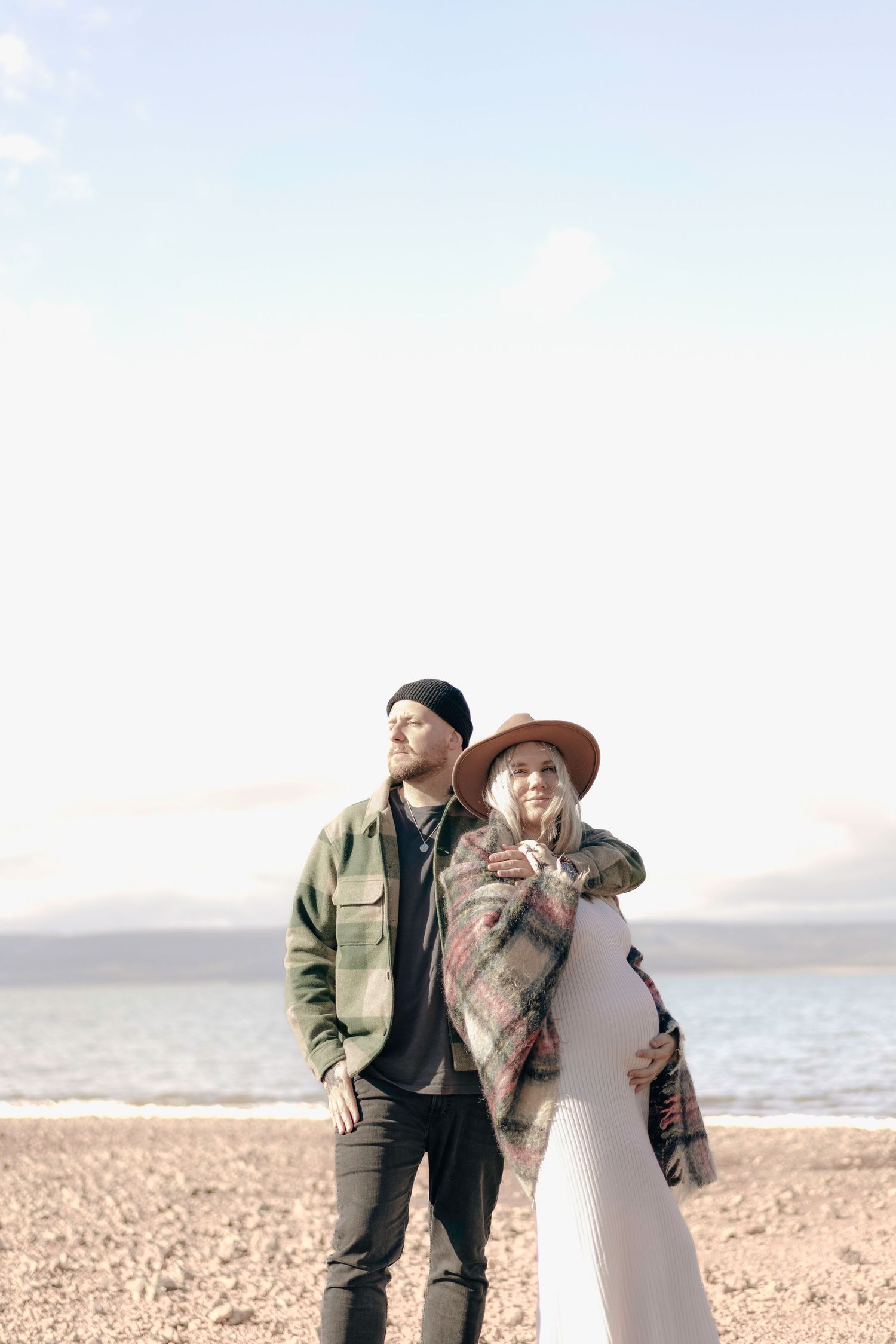 Pregnant couple standing on a sunny pebble beach, woman in boho hat and plaid wrap blanket.