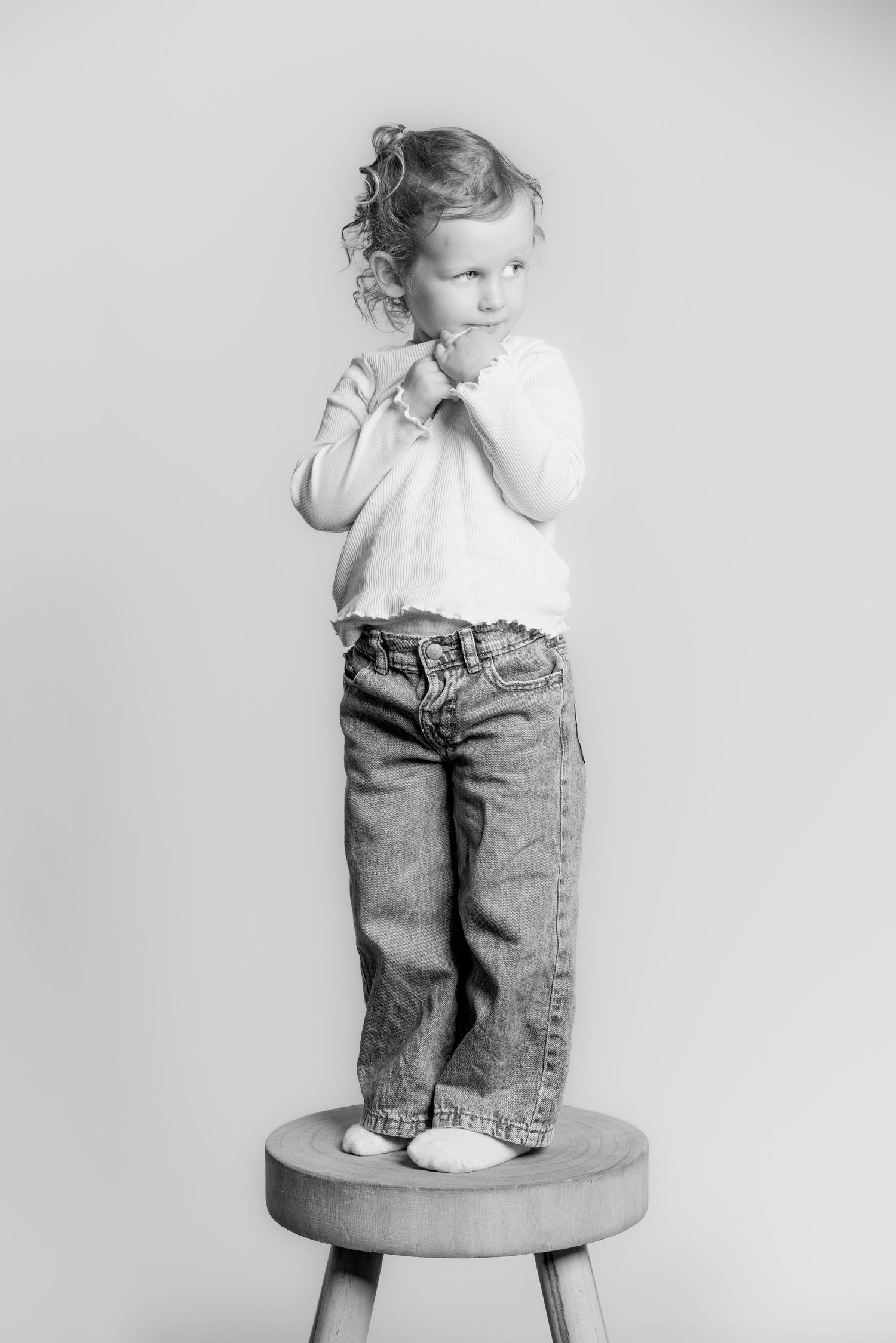 Black and white photo of a curly-haired girl standing on a stool in jeans and a white top.