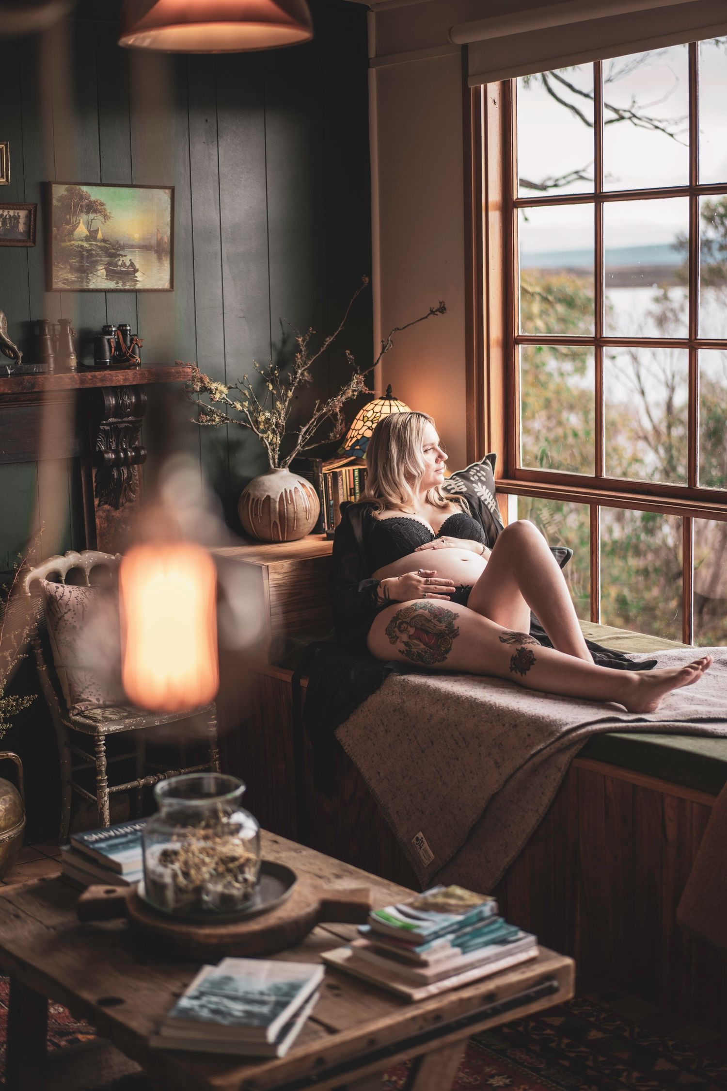 Tattooed pregnant woman relaxing on a couch by a large window in a cozy room.
