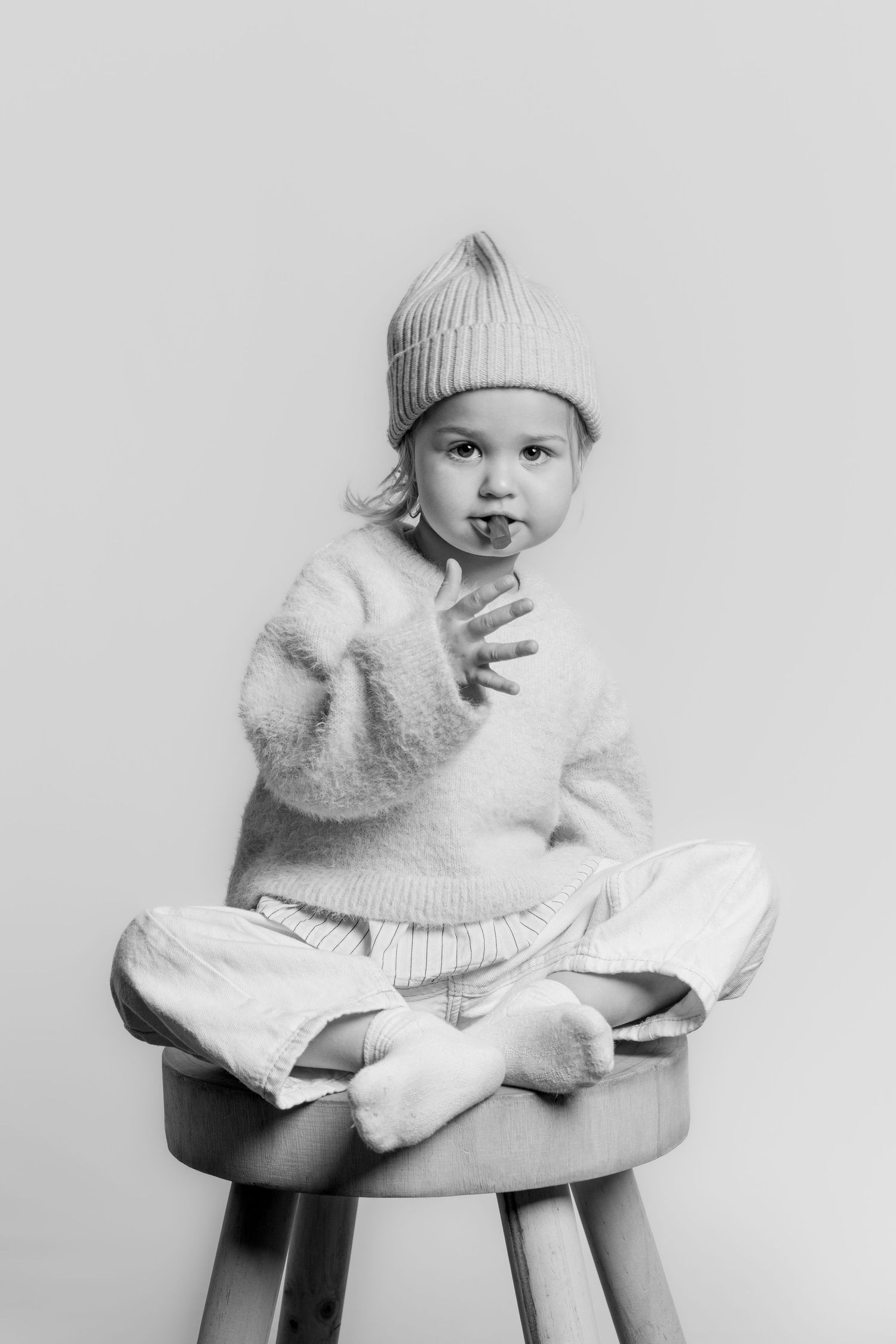 Black and white photo of a playful toddler in a knit beanie and sweater sitting cross-legged on a wooden stool.