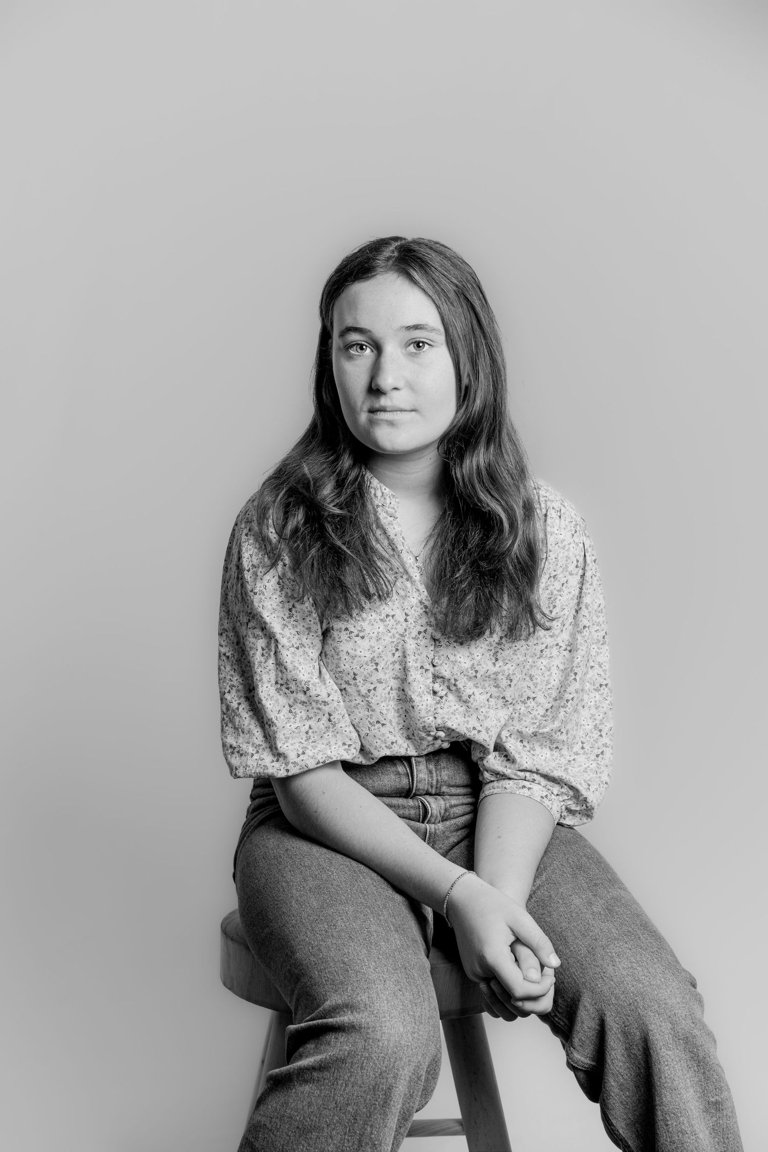 Black and white studio portrait of a teenage girl in a floral shirt sitting on a stool, calm expression.