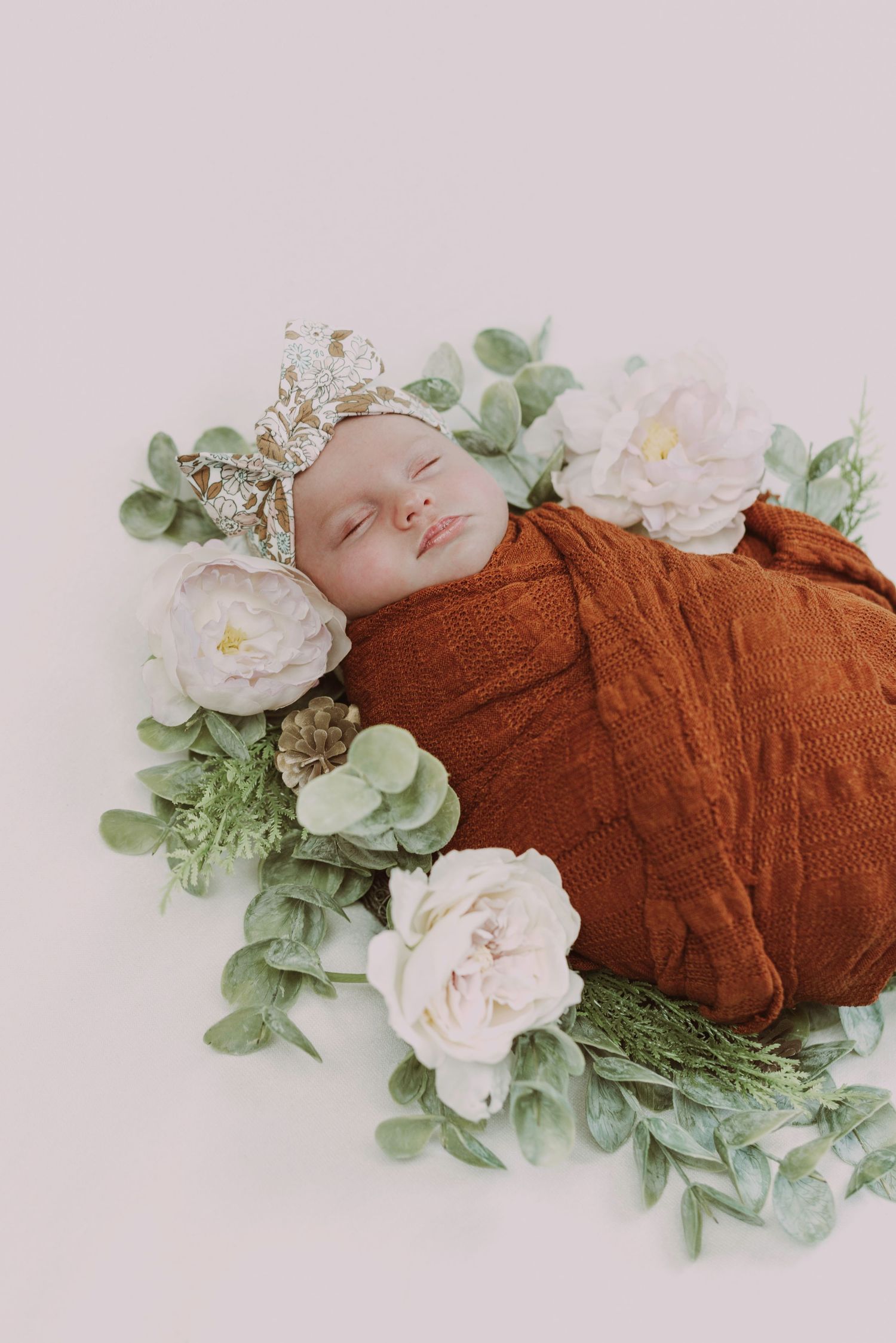 Peacefully sleeping newborn swaddled in rust orange wrap surrounded by white peonies and eucalyptus greenery.
