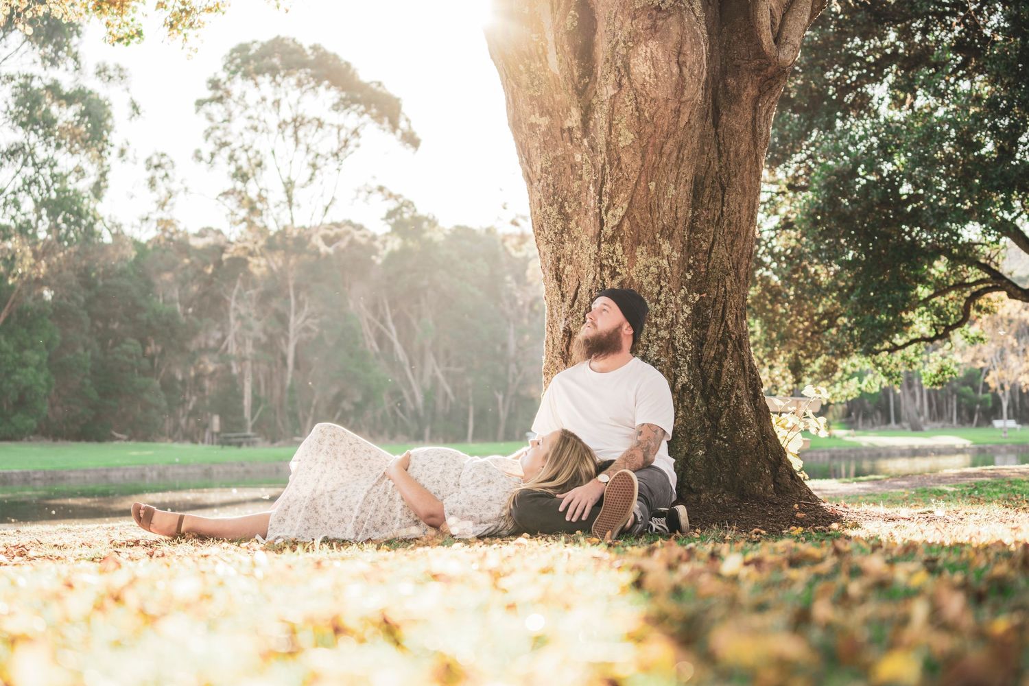 Bearded tattooed man and pregnant woman relaxing under a large tree in golden sunlight park.