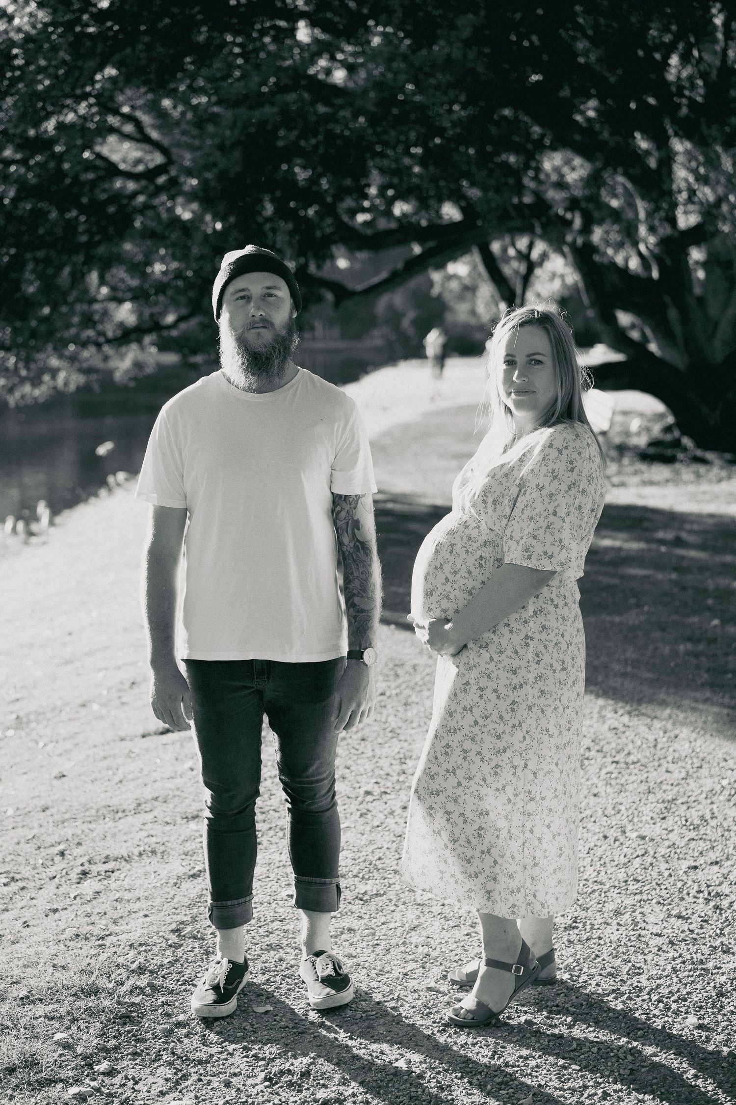 Tattooed bearded man and pregnant woman in floral dress posing outdoors, black and white photo.
