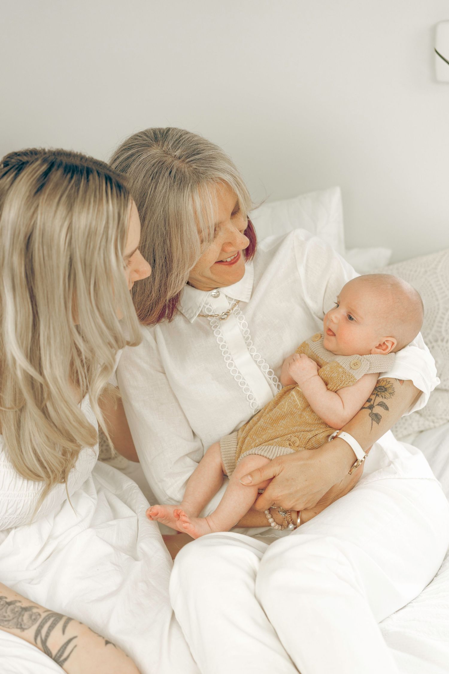Three generations bond as grandmother holds alert newborn baby while mother looks on in white bedroom setting.