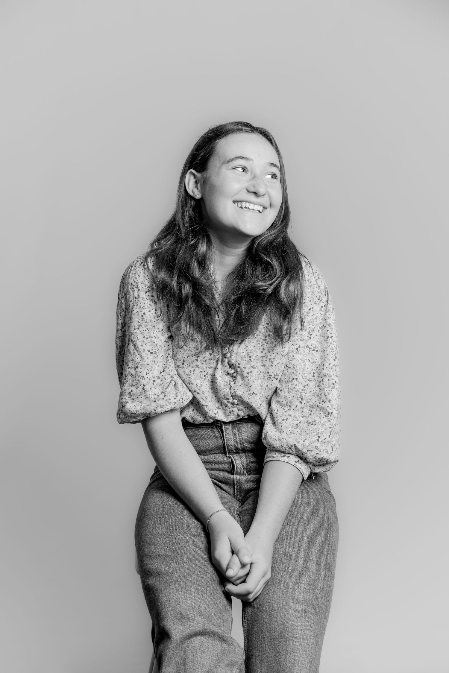 Black and white portrait of a smiling teenage girl with wavy hair sitting on a stool, hands clasped.