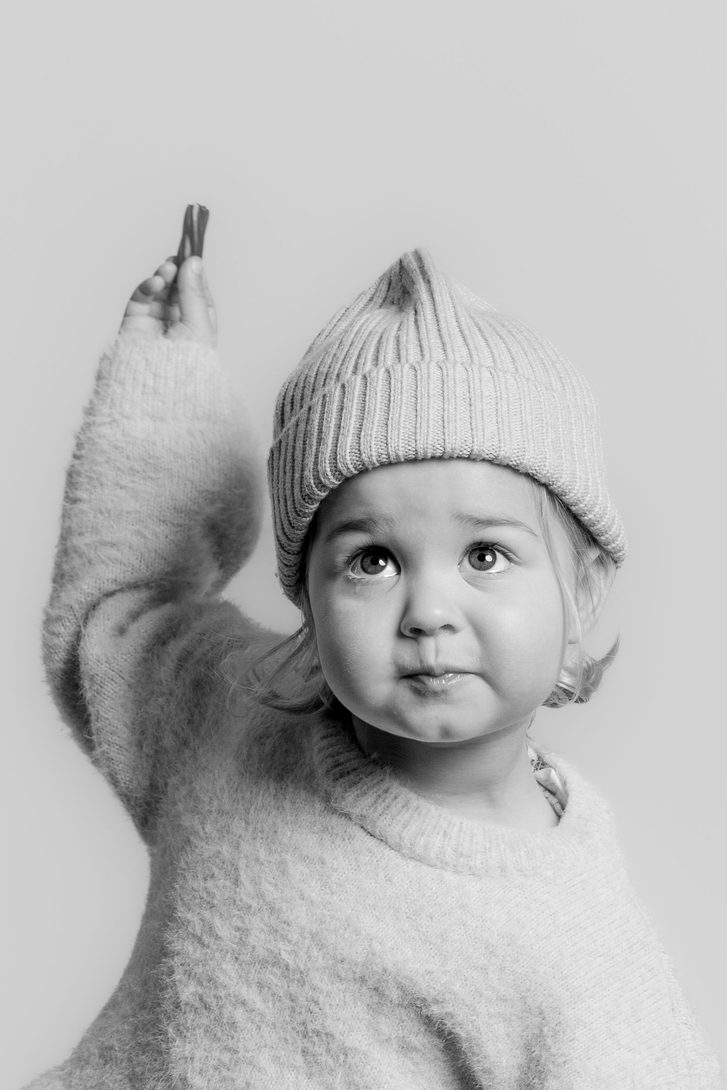 Black and white photo of a curious toddler in a knit hat and sweater raising their arm upward while making a face.