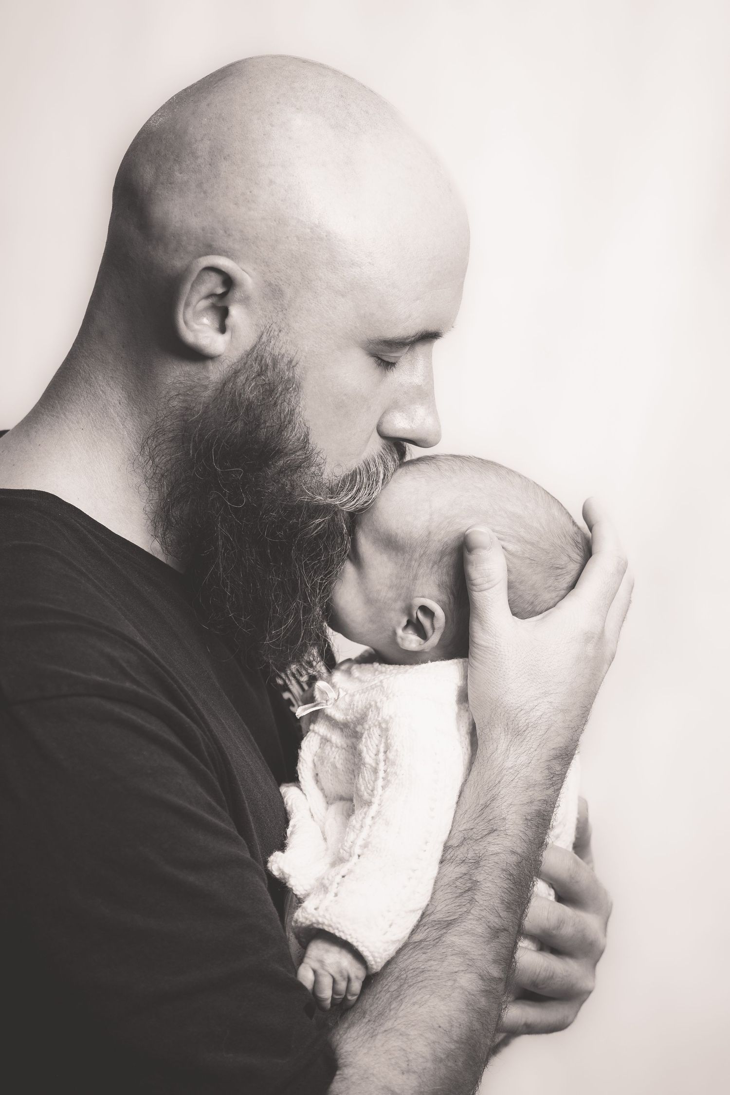 Bald bearded father gently kissing his sleeping newborn baby in black and white photo.