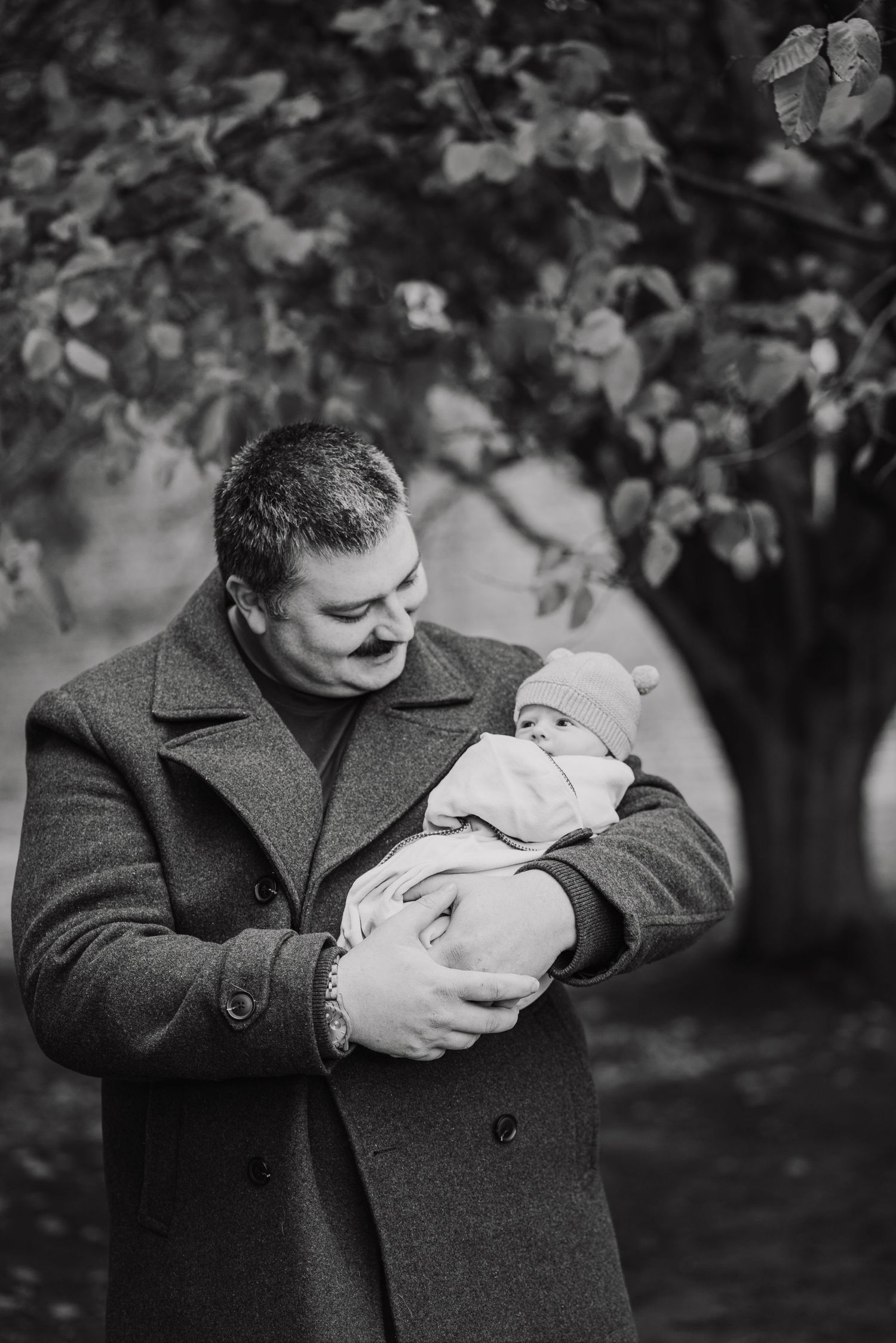 Black and white photo of father in coat tenderly holding newborn baby in park.