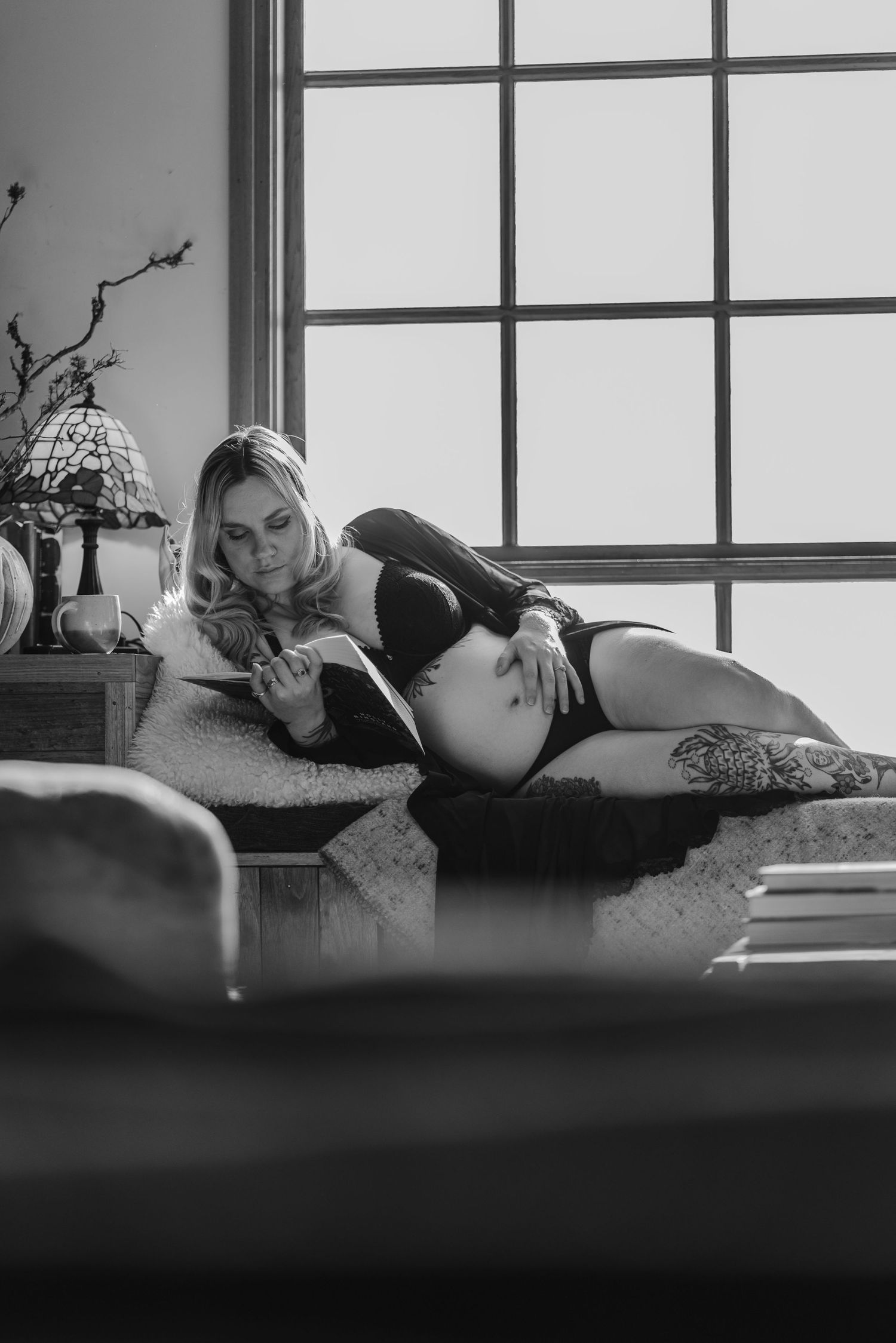 Black and white photo of a tattooed pregnant woman reading while lying by a window.