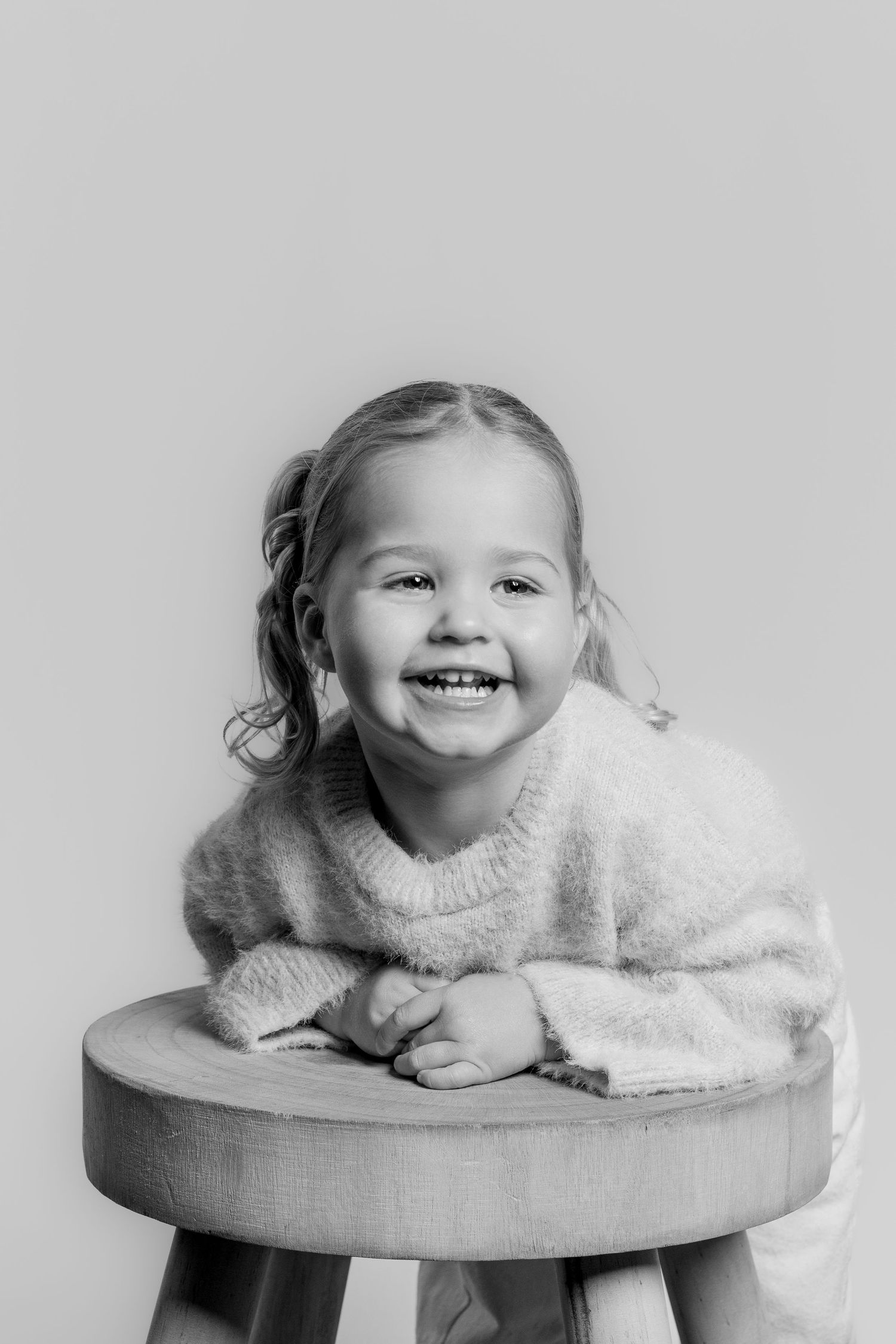 Black and white portrait of a laughing young girl leaning on a wooden stool with a big smile.
