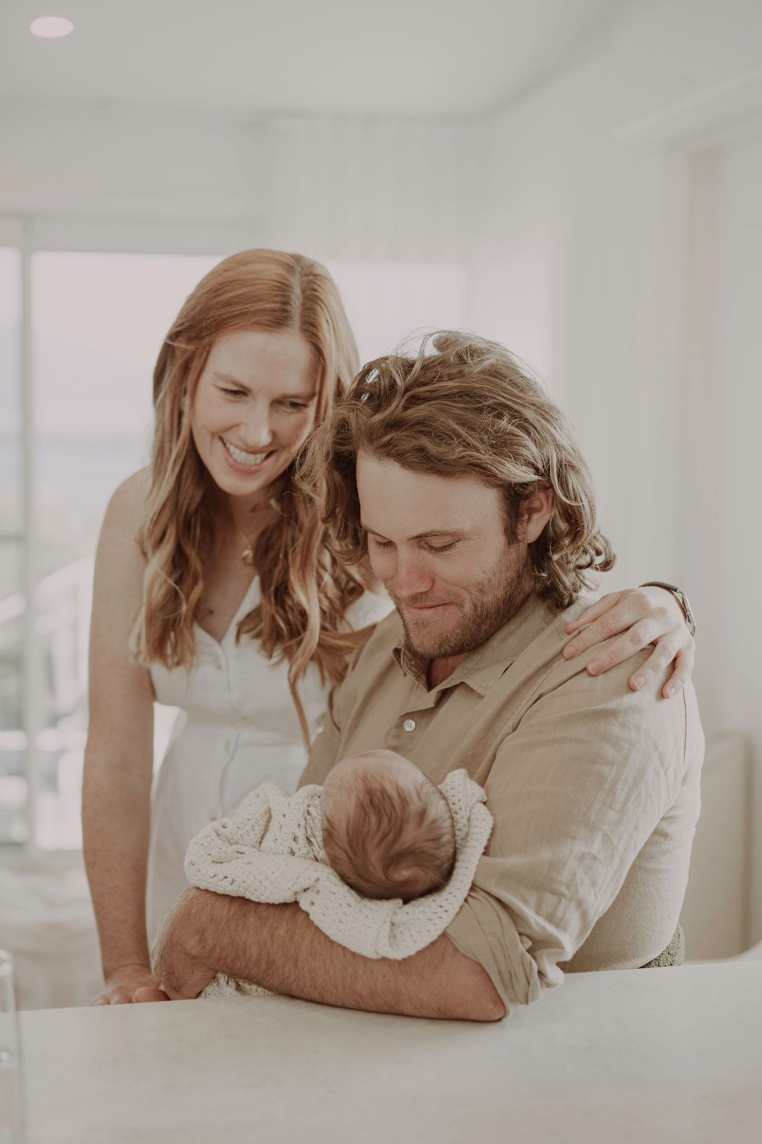 Smiling new parents admire their newborn baby in a bright white modern home setting.