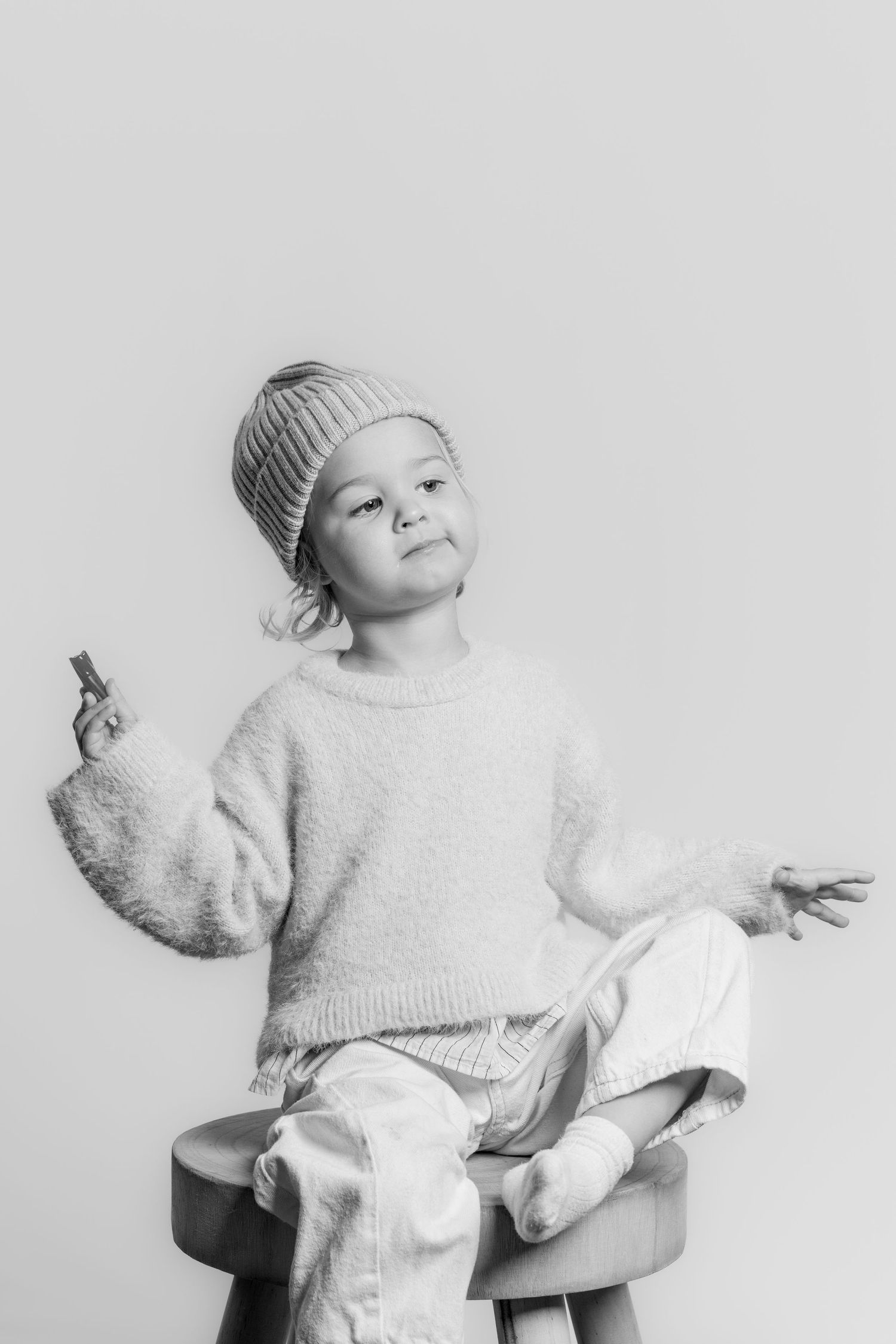 Black and white studio portrait of a child in a beanie hat sitting on a stool with arms outstretched.