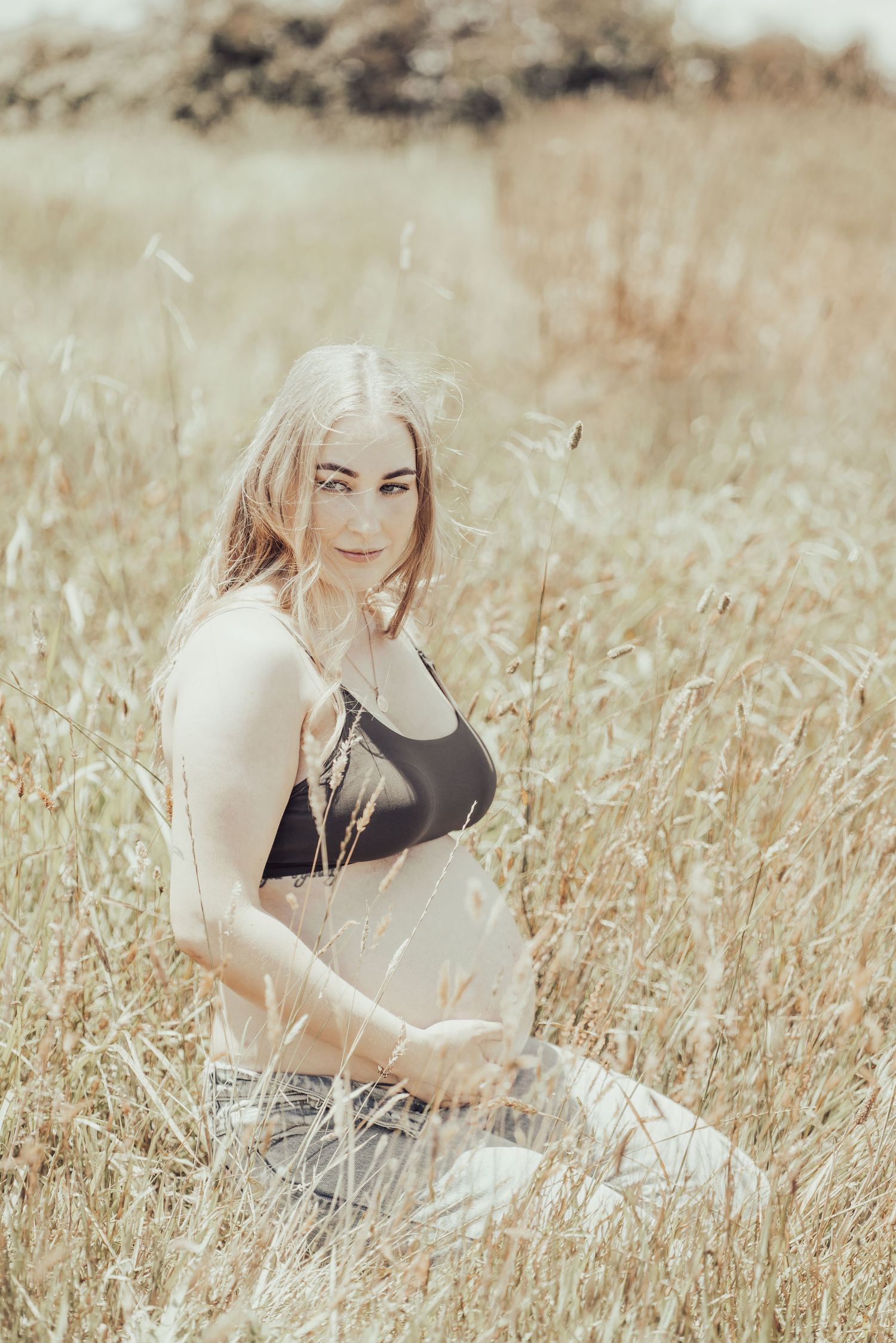 Pregnant woman in black crop top sitting in tall golden grass field during outdoor maternity shoot.