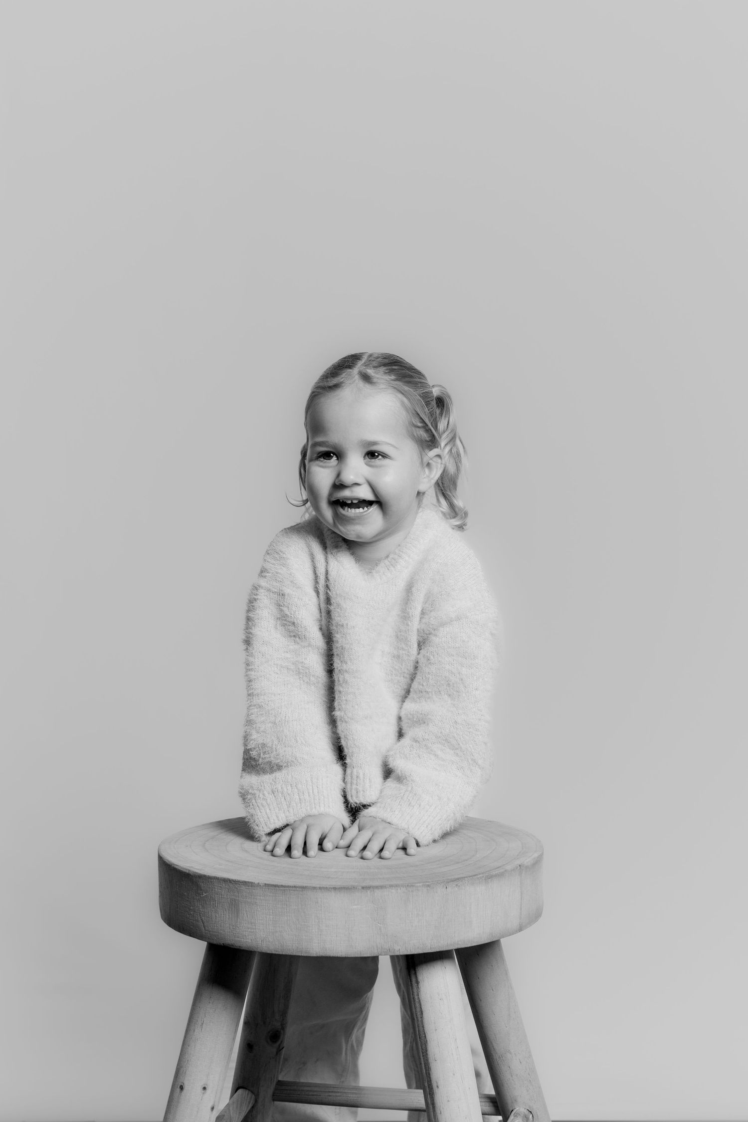 Black and white portrait of a laughing young girl sitting on a wooden stool, wearing a fluffy sweater.