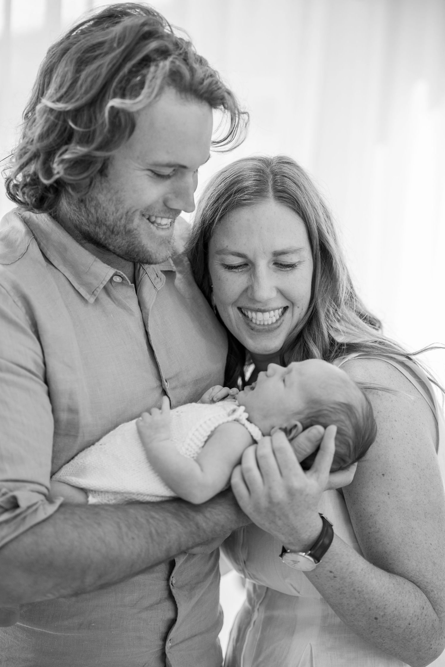 Joyful new parents laugh together while holding their swaddled newborn baby in black and white photo.