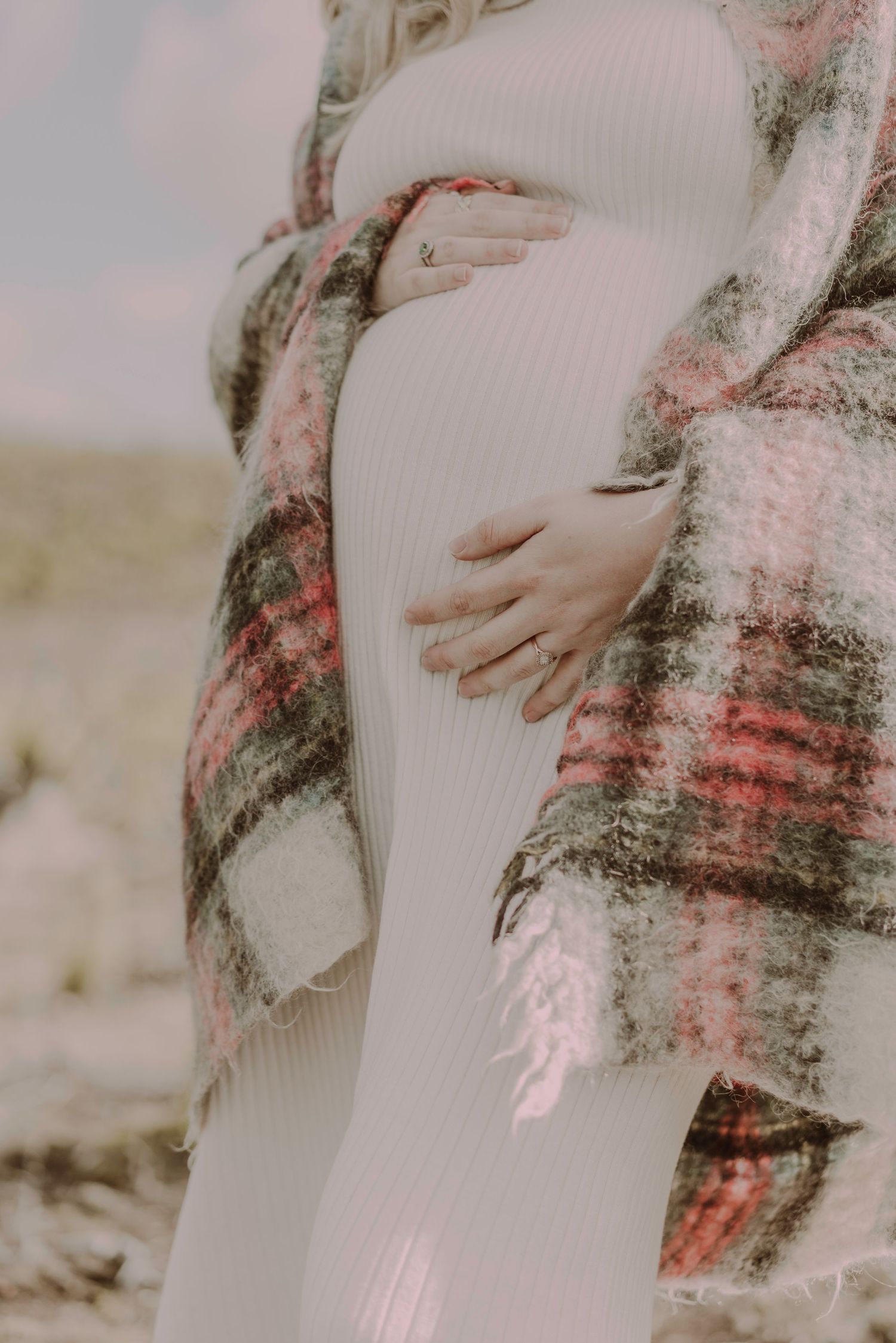 Close-up of pregnant woman in white ribbed dress holding baby bump wrapped in plaid blanket outdoors.