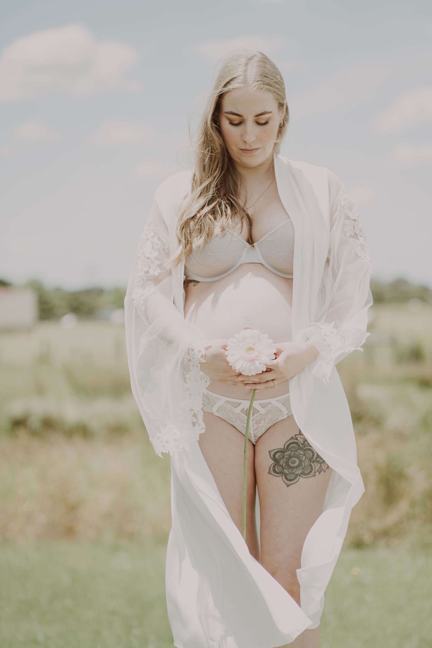 Pregnant blonde woman in white lace lingerie and robe holding flower in sunny meadow.