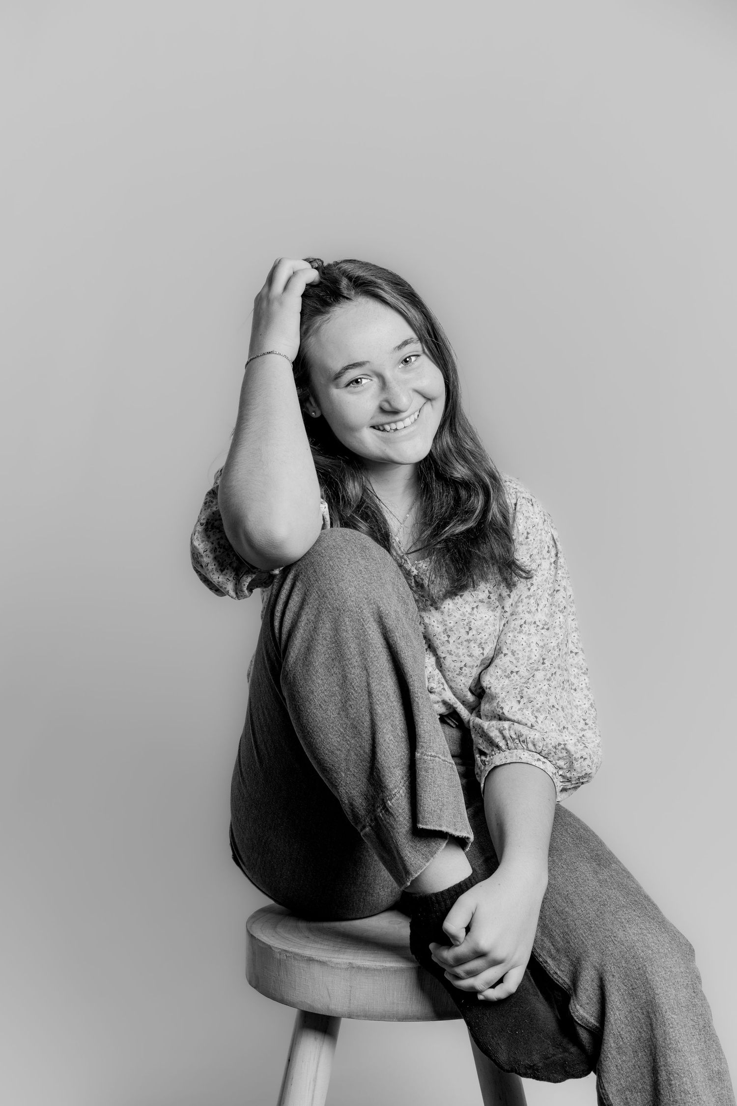 Black and white studio portrait of a teenage girl sitting on a stool, hand in hair, smiling.