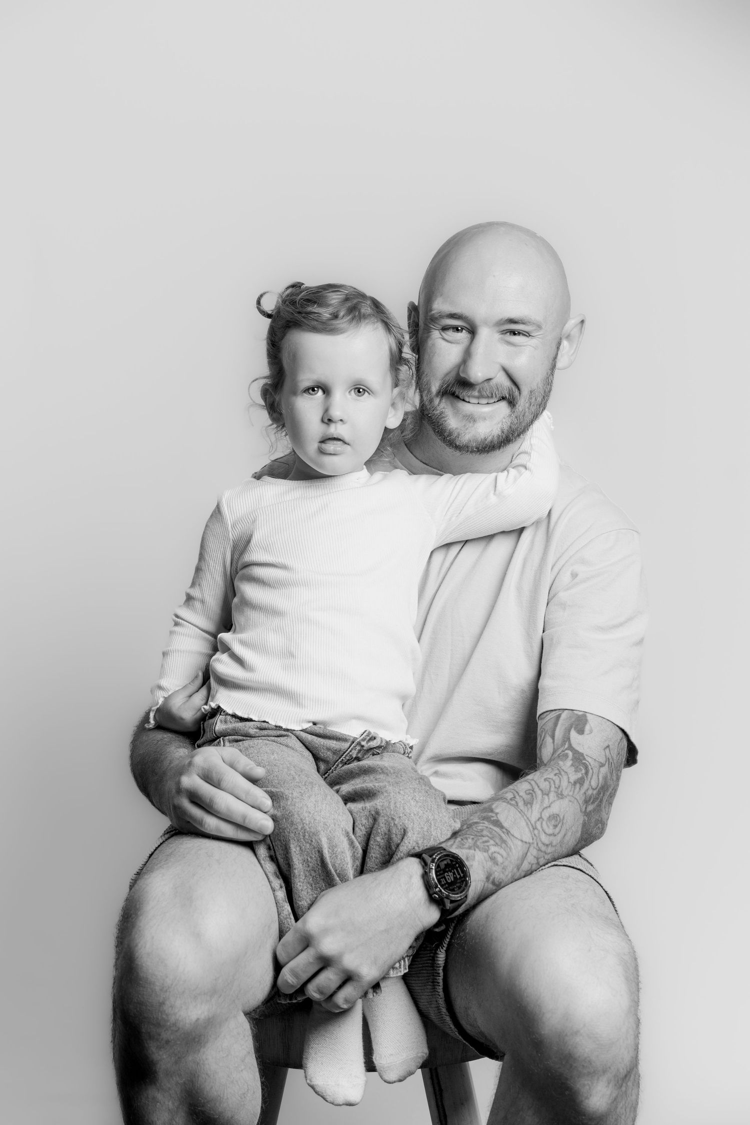 Black and white studio portrait of a tattooed father sitting with his young daughter on his lap.
