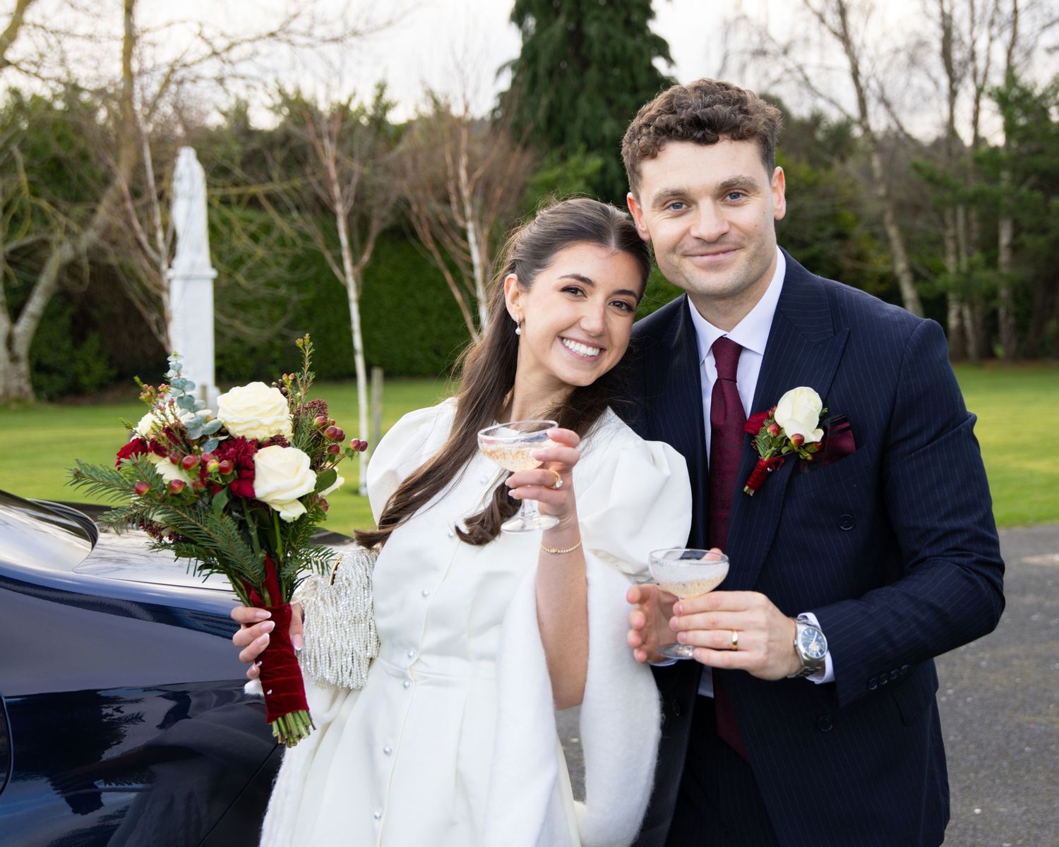 Happy newlyweds pose outdoors, bride holding bouquet and groom in navy suit with boutonniere, both holding champagne glasses.