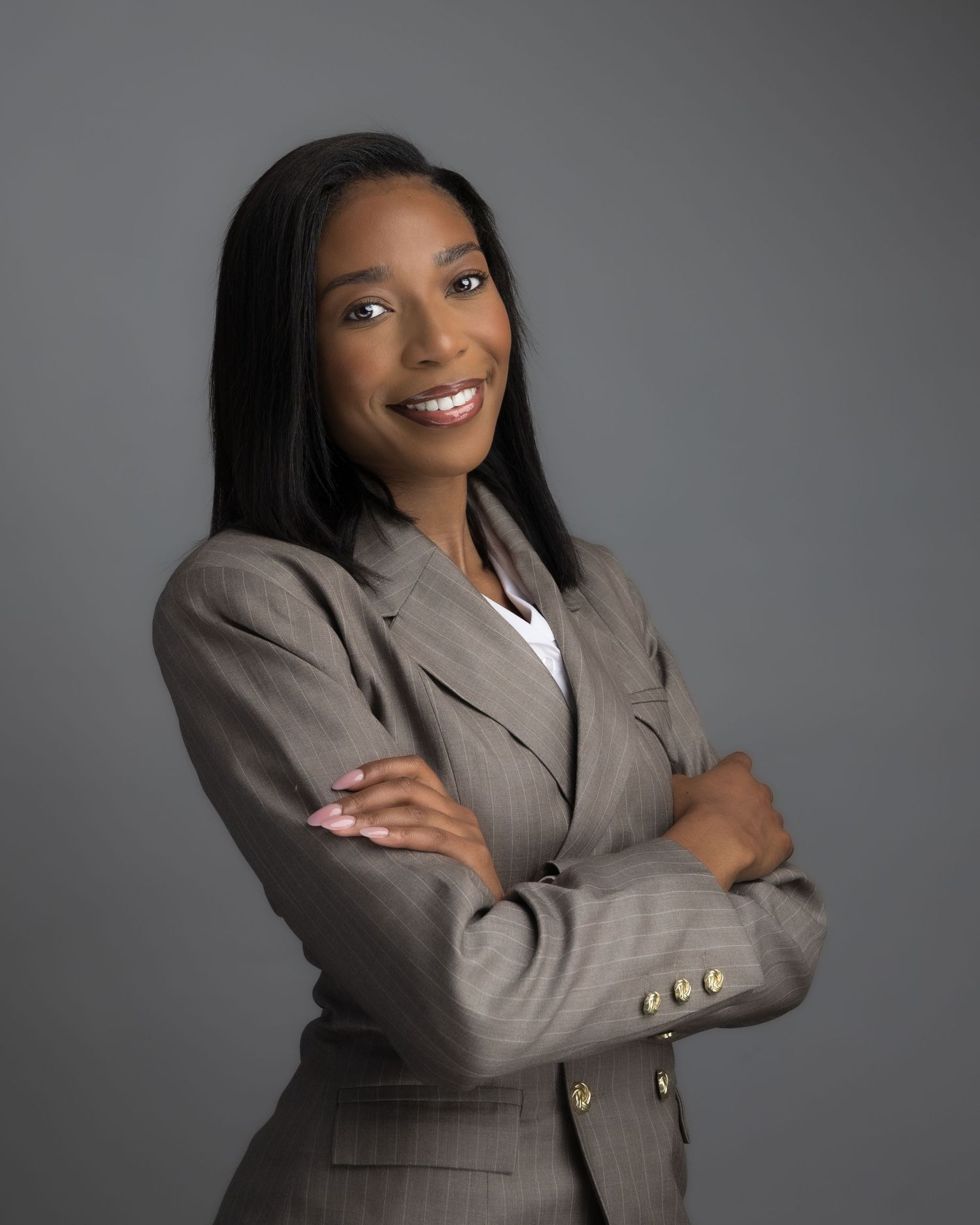 Professional woman in gray blazer smiling with arms crossed against gray background.