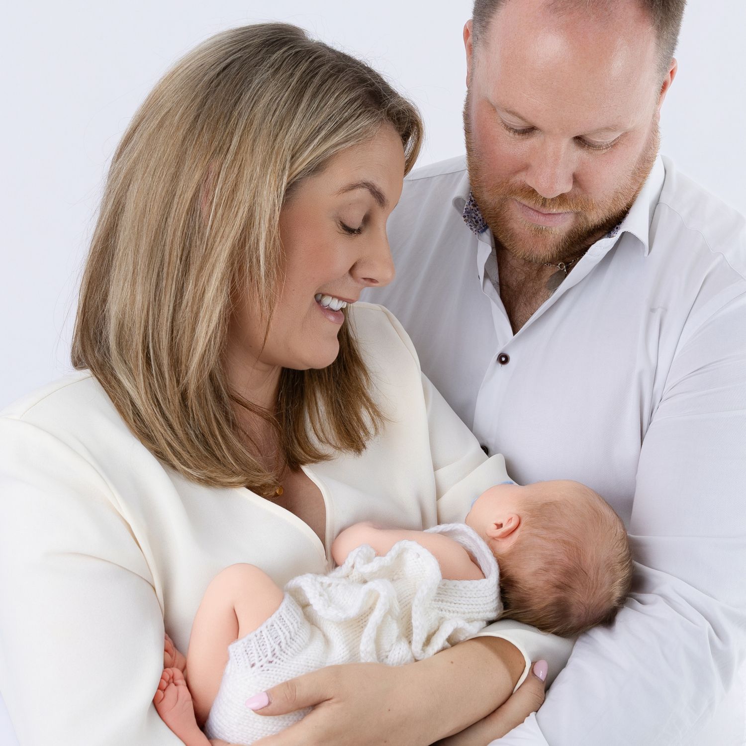 A couple in white clothing lovingly gazing at their newborn baby against a white background.